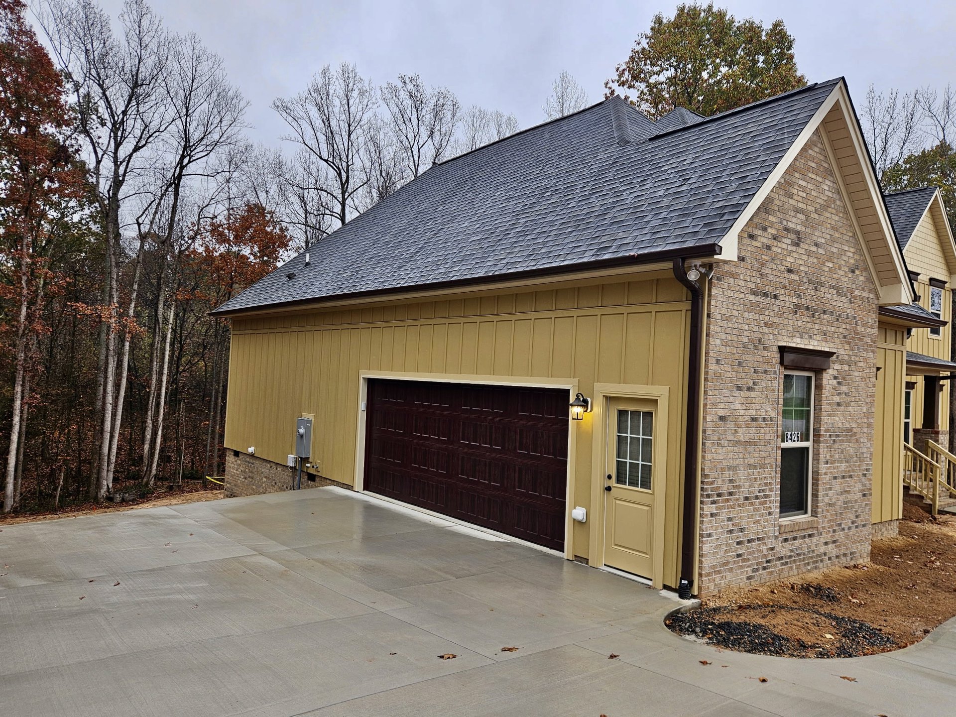 Gray garage door with white trim on a modern home, surrounded by trees with brown leaves, large windows, and a dark shingle roof.