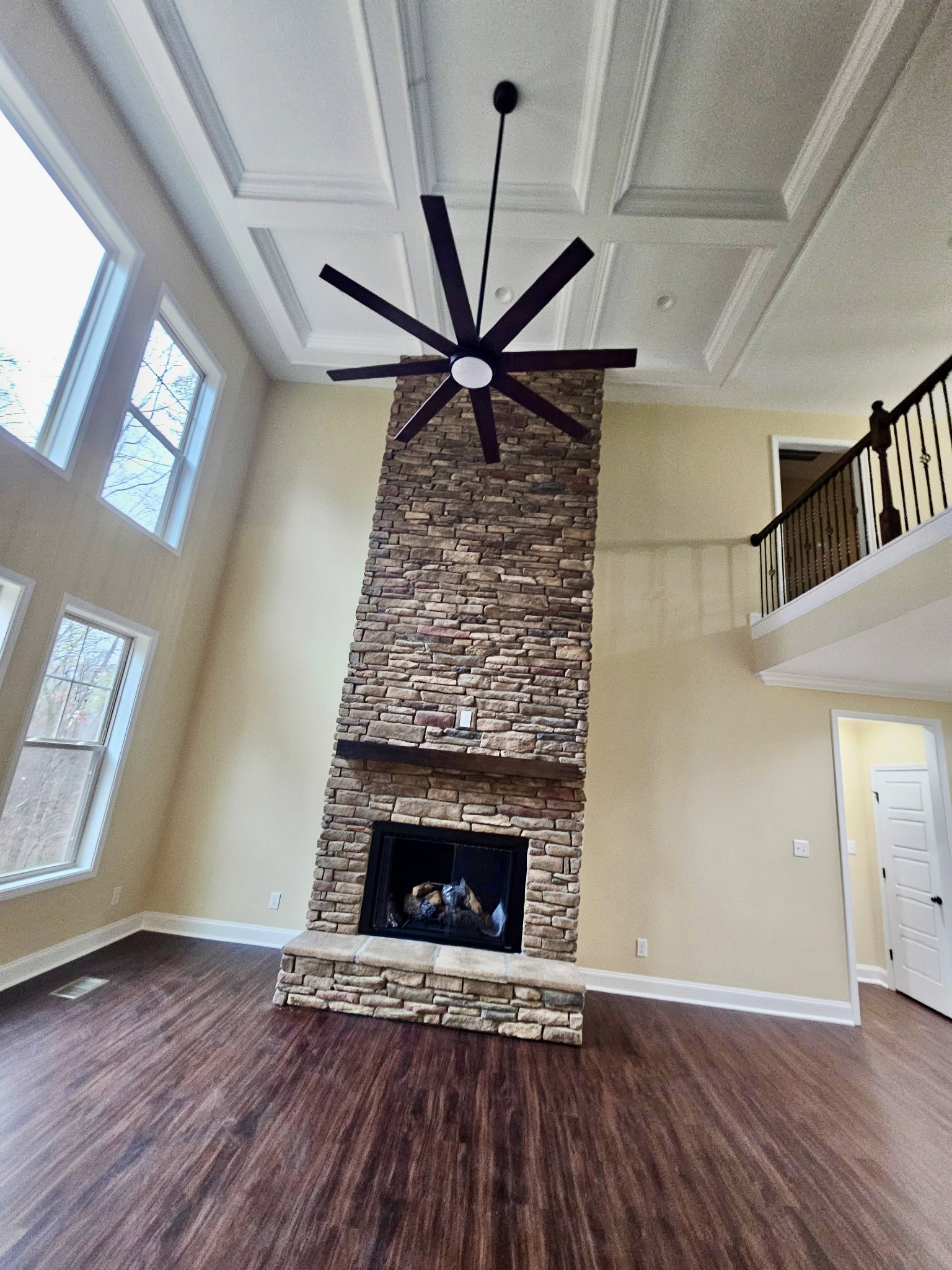 Living room with brick fireplace featuring black frame, hardwood flooring, ceiling fan with light, white molding, and staircase with black railing