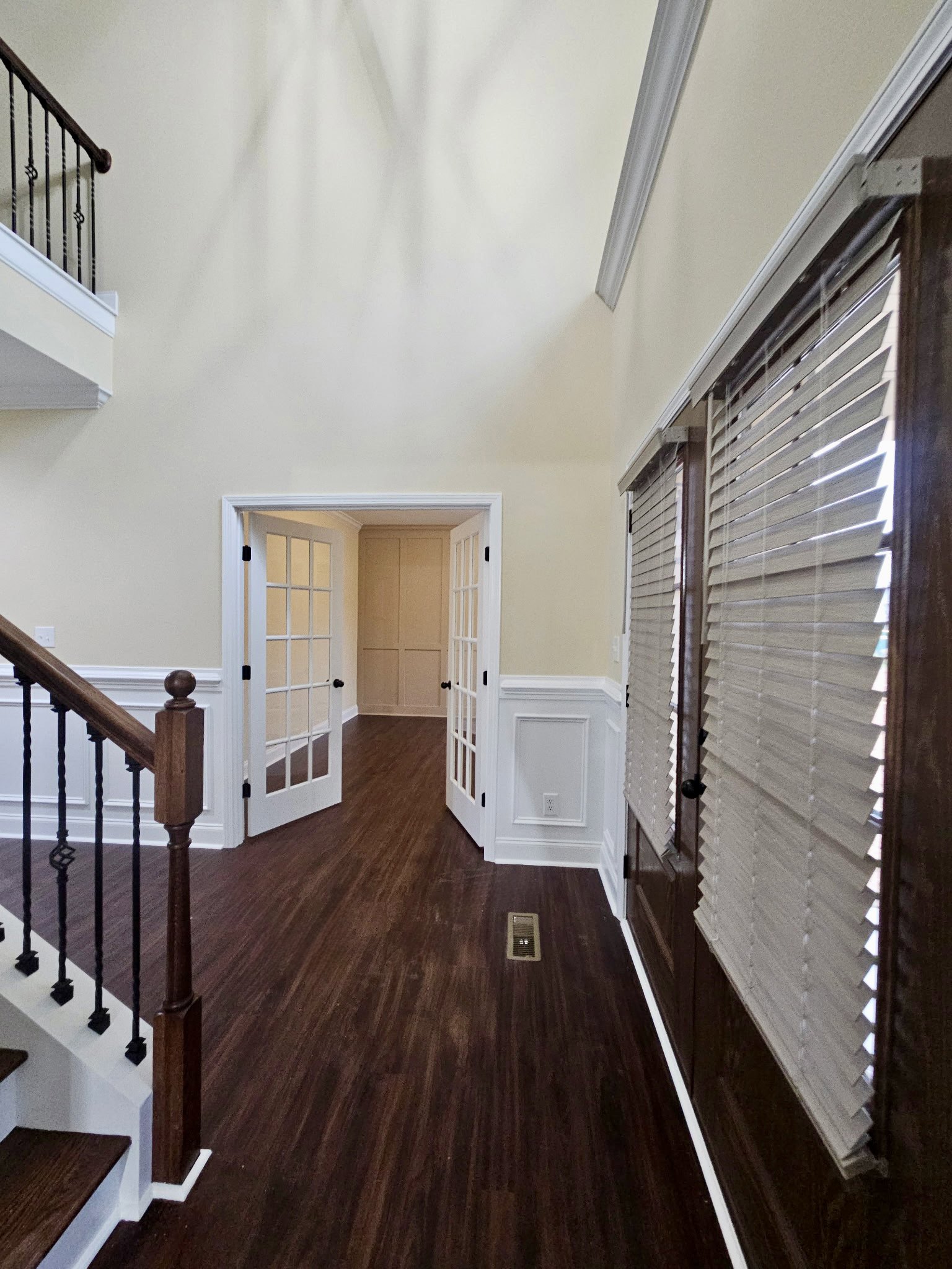 Hallway with hardwood floors, white walls, wooden staircase with metal handrail, glass-paneled door, window with blinds and vent