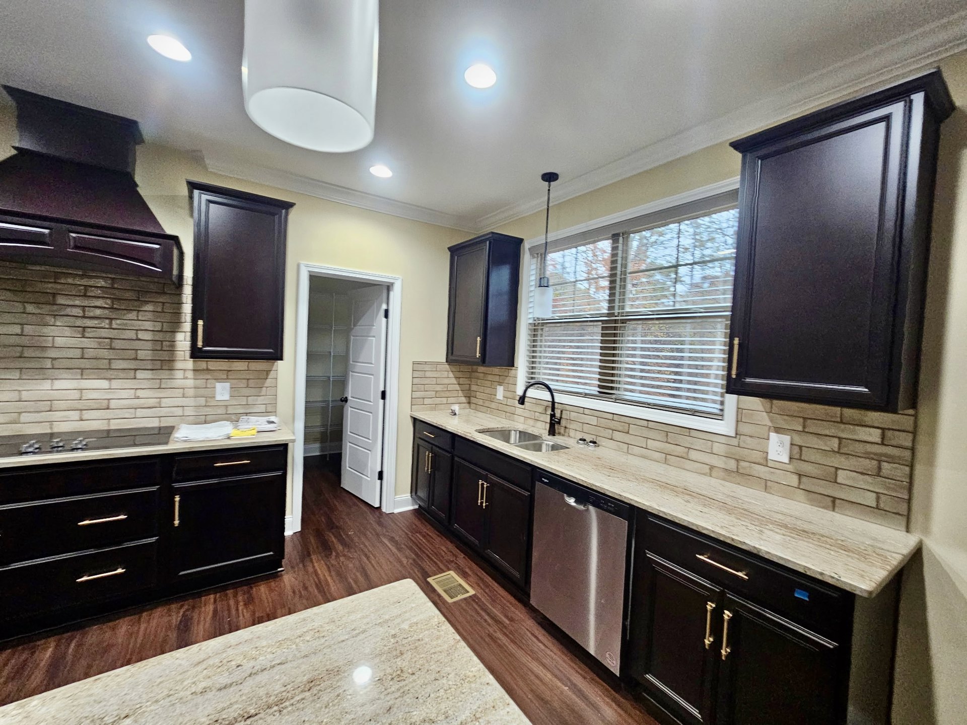 Kitchen with dark wood cabinets, granite countertops, stainless steel sink, wood flooring, white door with black handle, black door with gold knob, and recessed ceiling light.