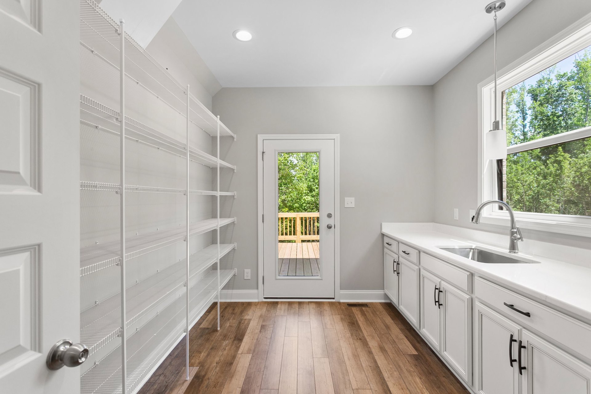White kitchen with empty white shelves, wood flooring, white cabinetry, and a door opening to a deck with trees visible outside.