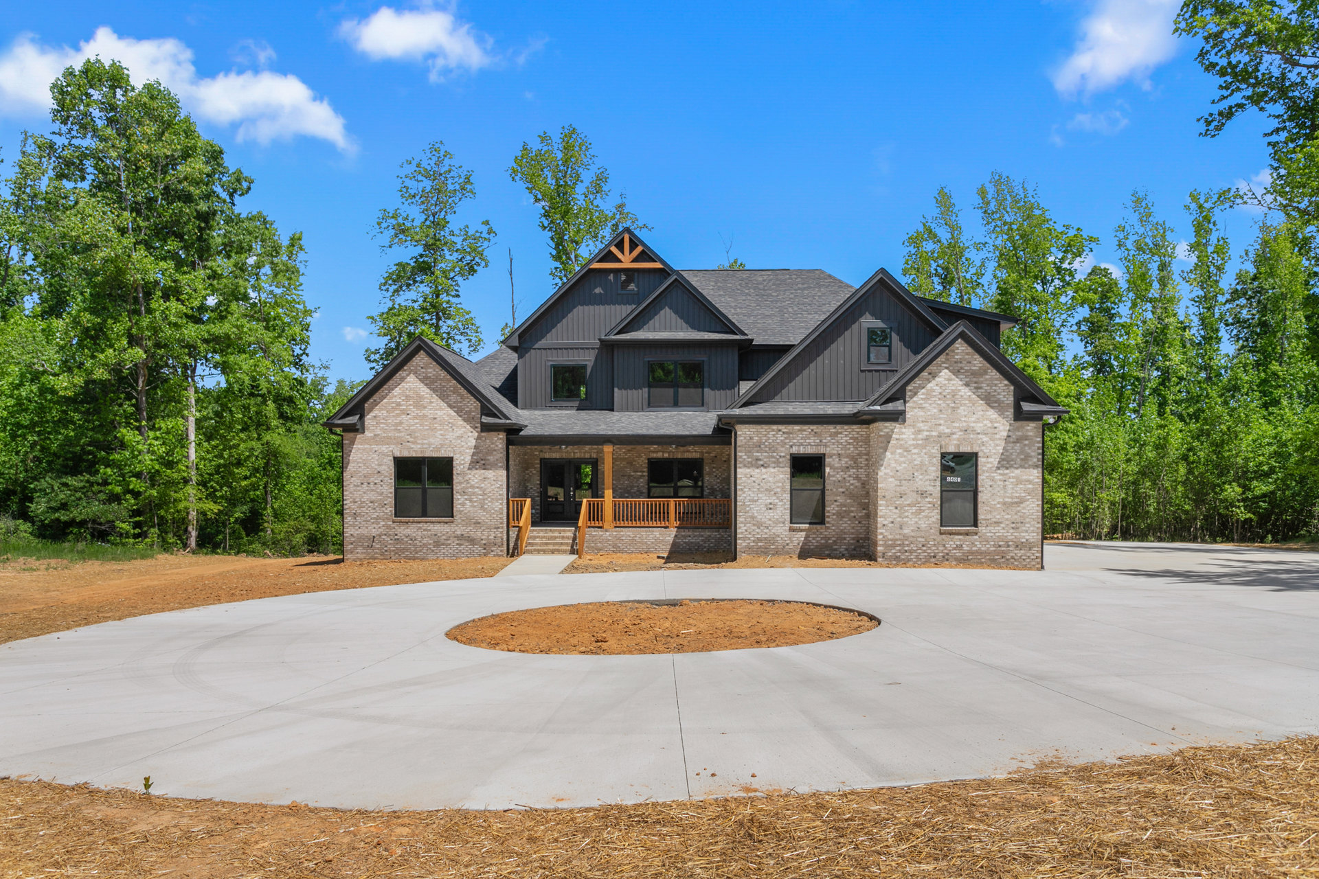 Two-story home with light siding, covered front porch, and circular concrete driveway bordered by dirt and landscaping, mature trees in background, screened window visible on side.