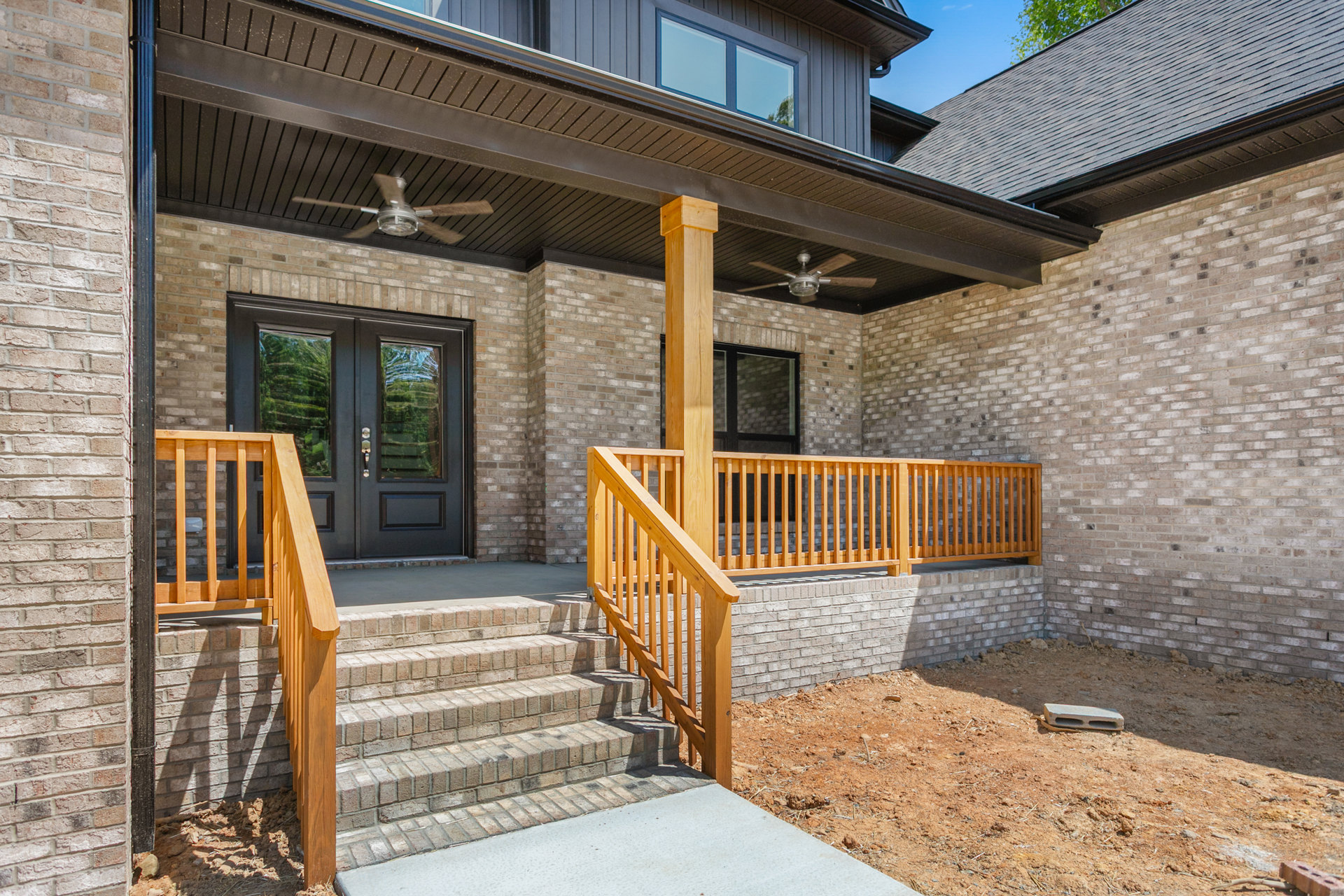 Red brick house with black double front doors featuring glass panes, wooden railing along porch stairs, and brick wall backdrop