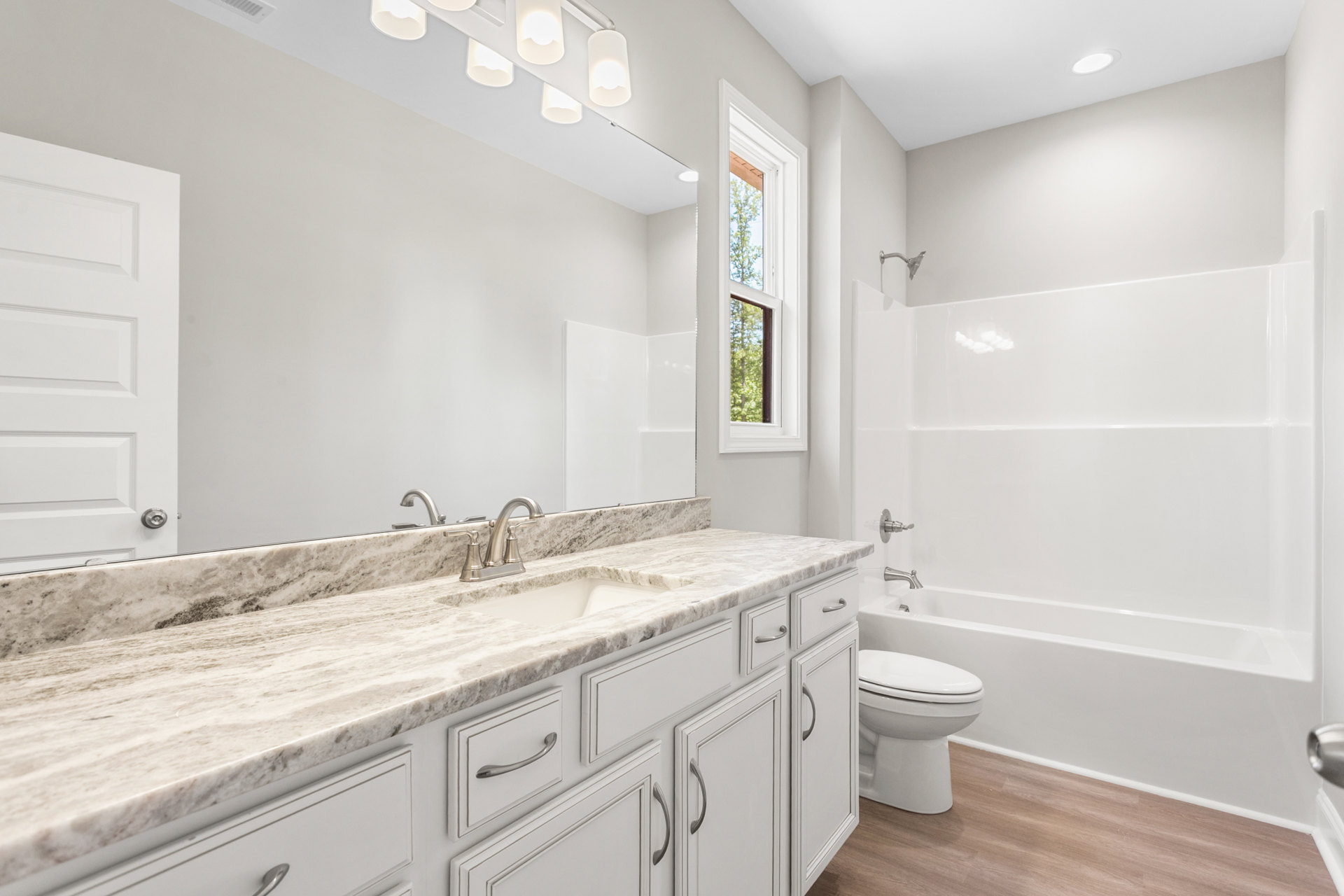 Bathroom with marble countertop, undermount sink, white toilet, wood cabinetry, round light fixture, window showing tree outside, and tiled walls.