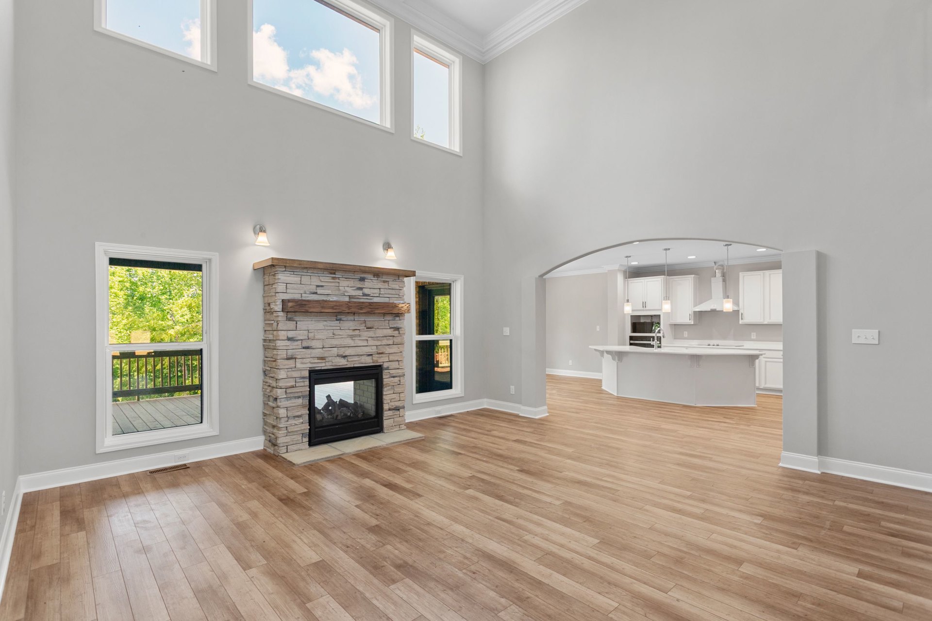 Spacious living room featuring hardwood floors, a fireplace with a glass door and wood mantel, open kitchen area, large window overlooking deck and trees, and skylight illuminating