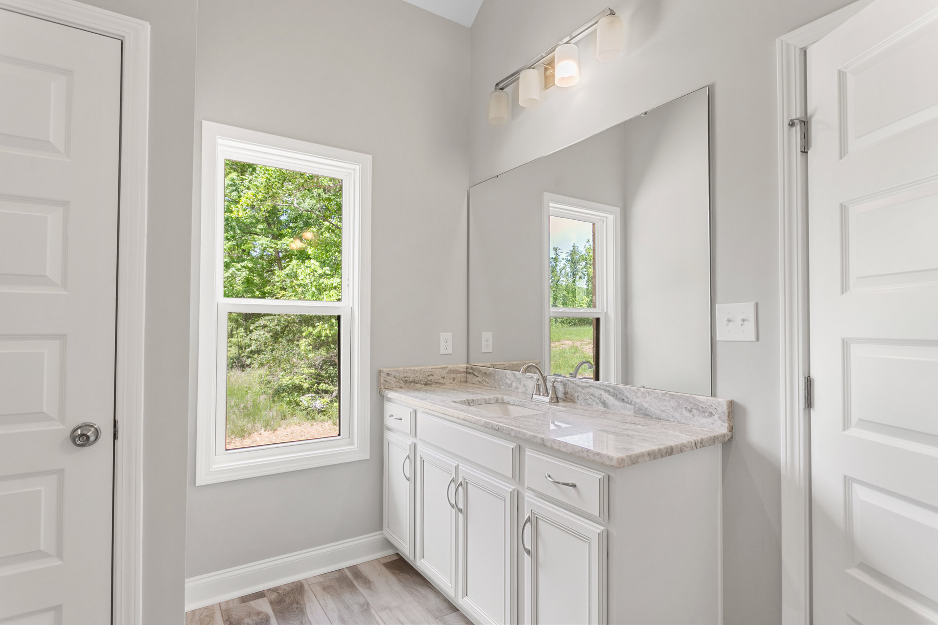 Bathroom featuring a marble countertop, undermount sink, large framed mirror, white cabinetry, and window overlooking trees