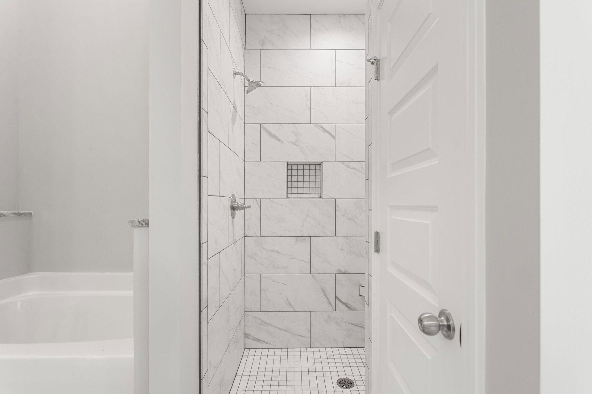 White tile shower with black grout, silver shower head, white door with silver knob, white tile floor with black lines