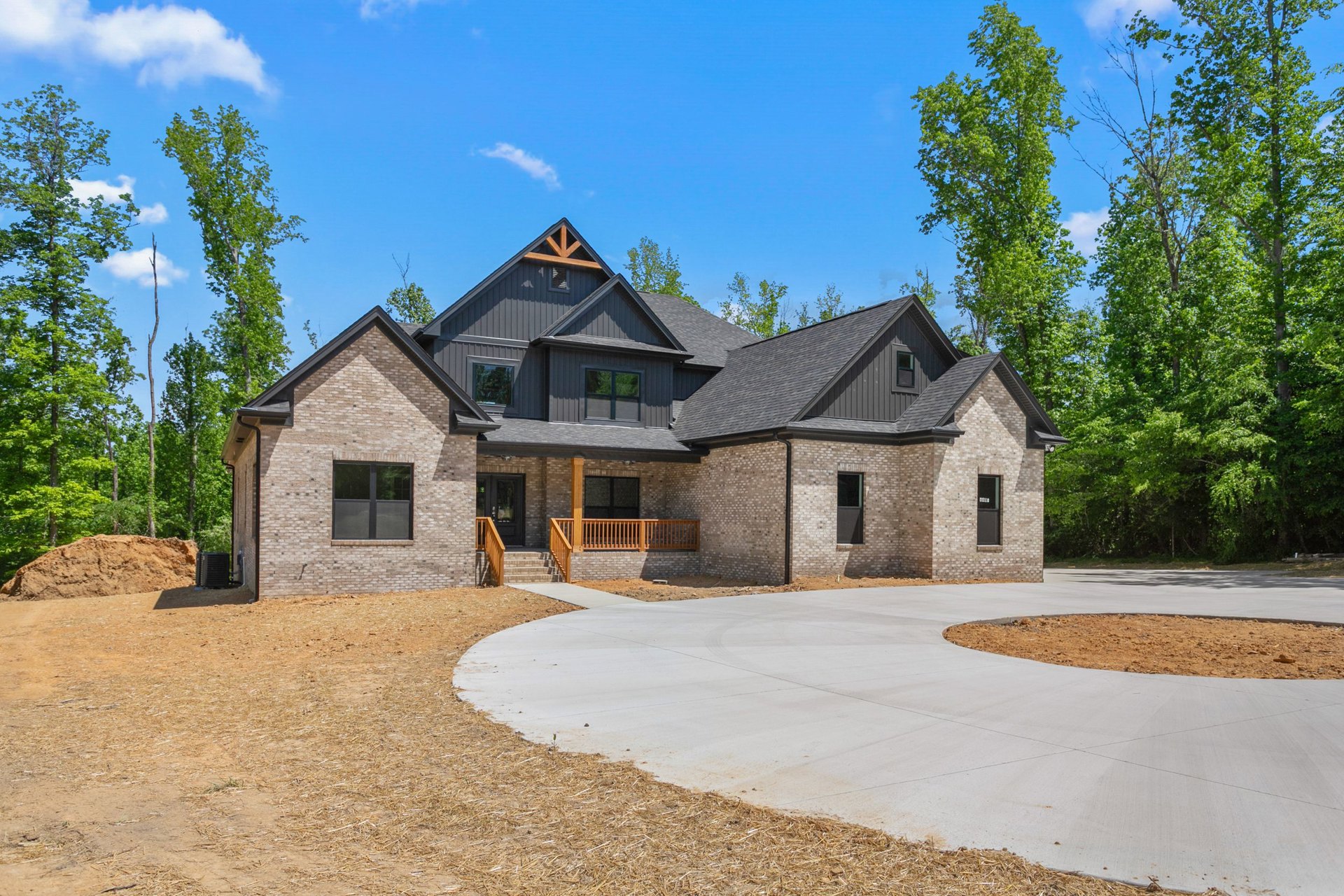 Two-story brick house with white trim, concrete driveway, mature trees in the background, wooden railing along front steps, large windows reflecting greenery, landscaped front yard