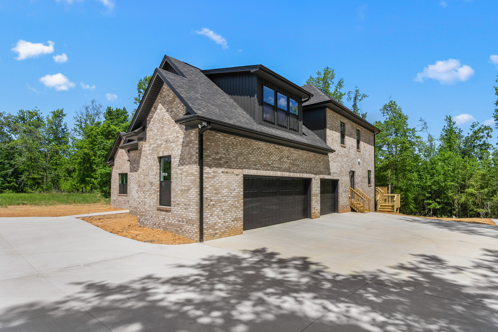 Brick house with attached garages, wooden deck and stairs, concrete driveway bordered by mature trees, man standing near garage door, sunlight casting shadows on lawn and pavement