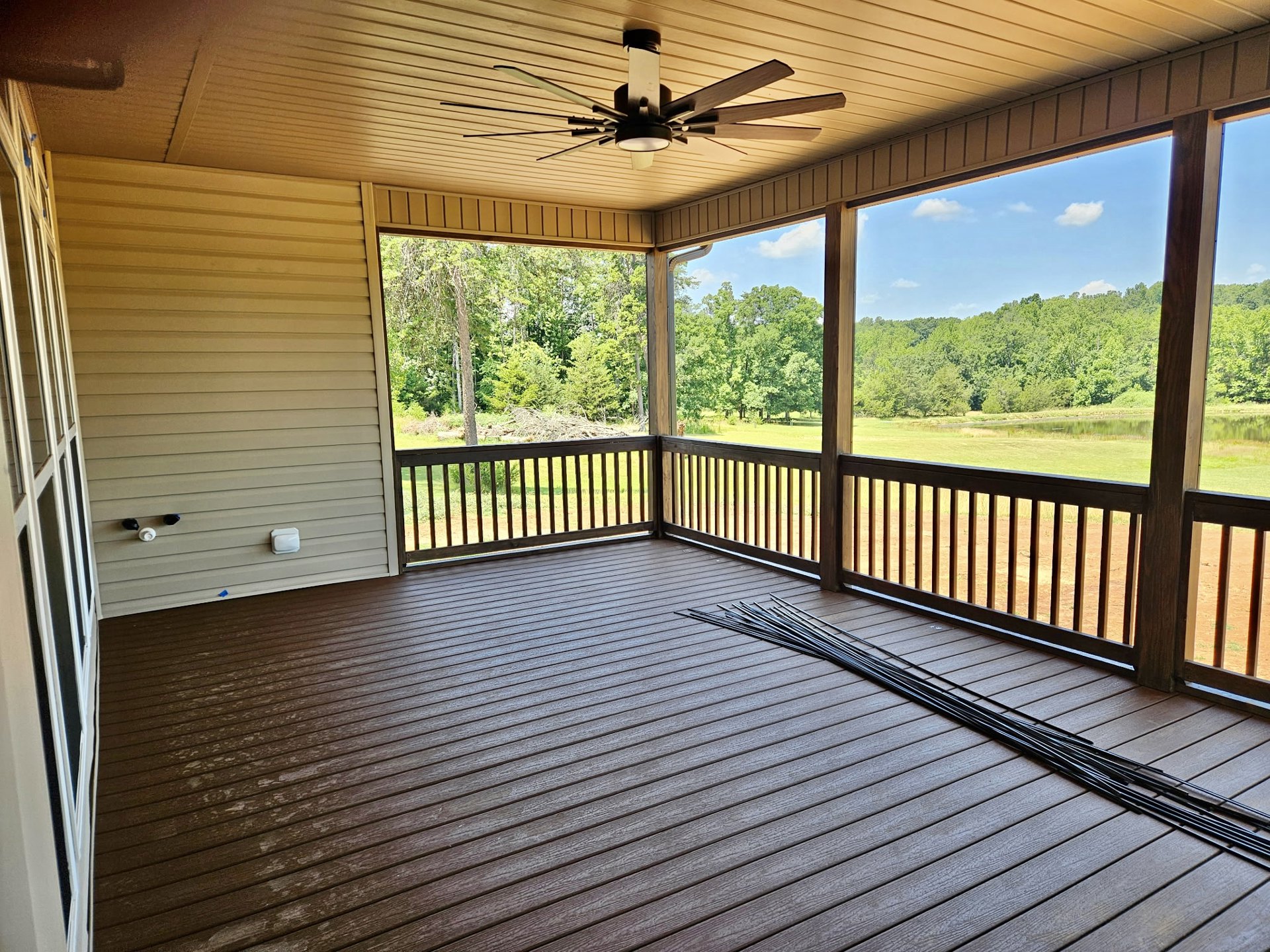 Wood deck with black metal railing, ceiling fan with light fixture mounted to covered ceiling, leafy trees visible beyond railing.