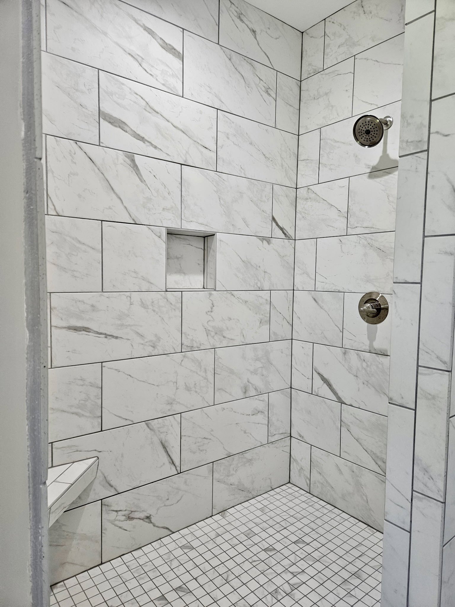 Shower with white tile walls and black border, round shower head, white tile floor with black and white accents, marble frame, close-up of metal knob
