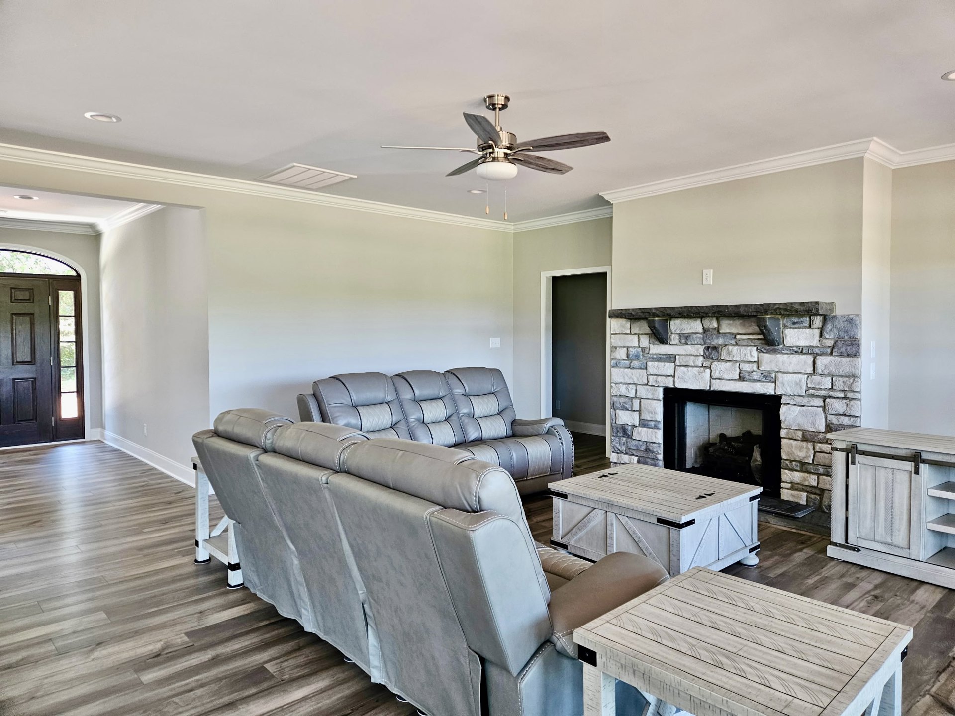 Living room with stone fireplace, two grey and white upholstered couches, wood coffee table, ceiling fan with light, neutral walls, hardwood flooring, and white built-in storage