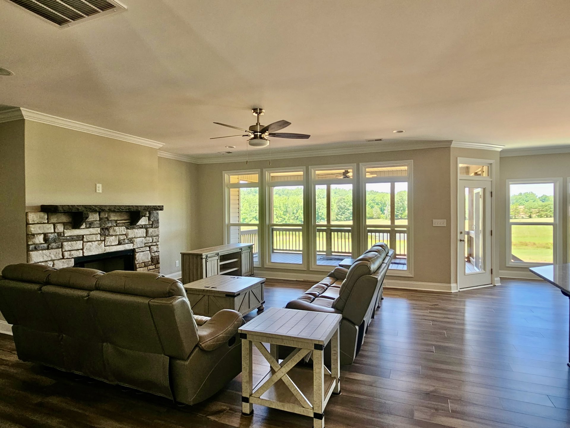 Stone fireplace with black frame, gray couches, white table with black metal legs, ceiling fan with light, vent in white ceiling, hardwood flooring, large windows, neutral walls