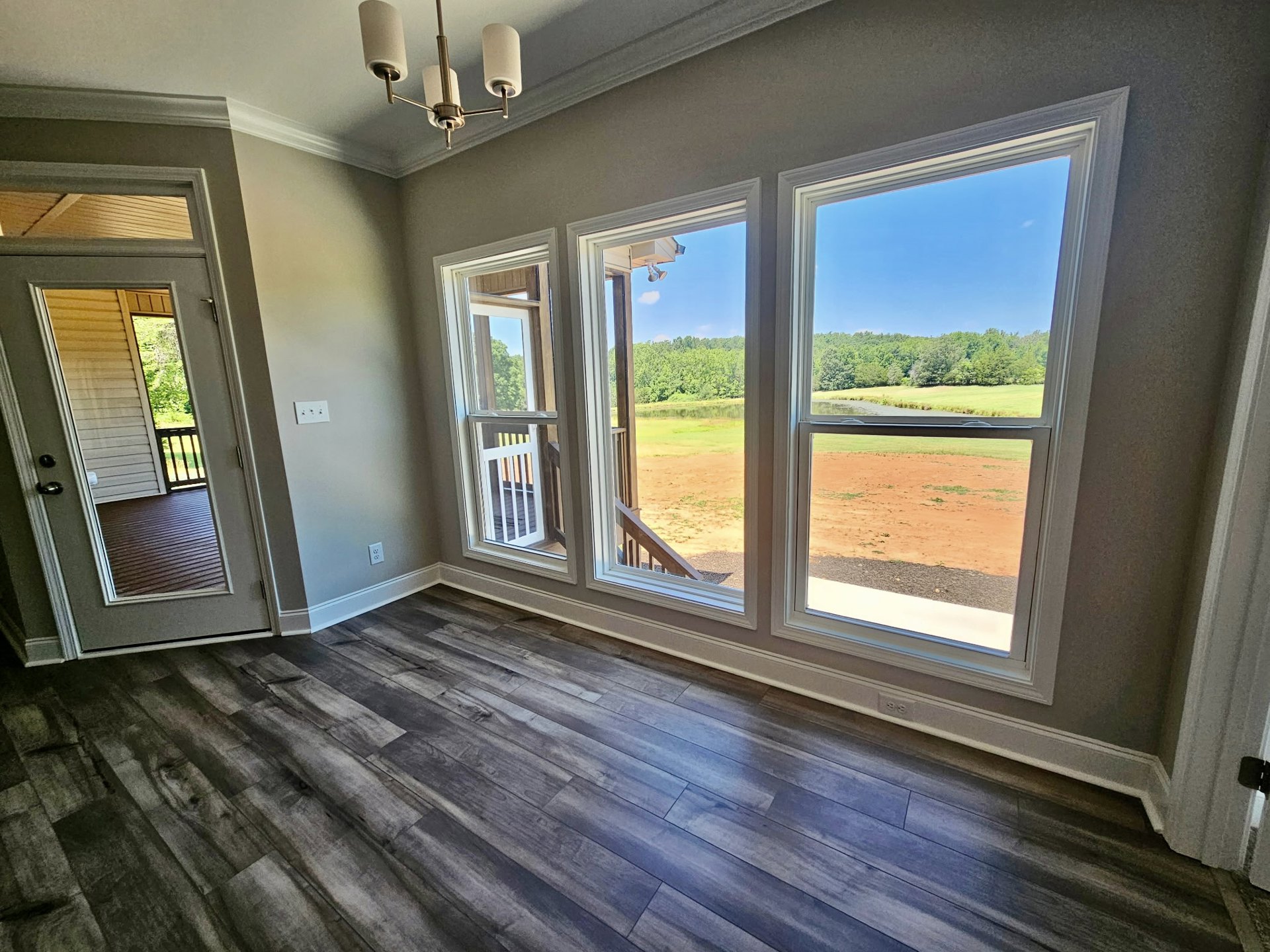 Open-concept living area with wood flooring, large windows overlooking a field and trees, modern chandelier, white walls, and staircase adjacent to entry door.