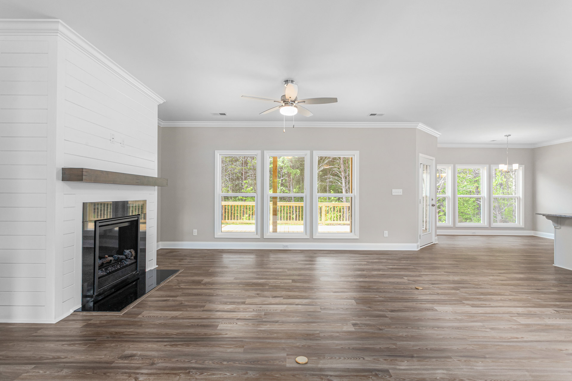 Living room with wood flooring, central fireplace containing logs, ceiling fan with light, large window showing trees outside
