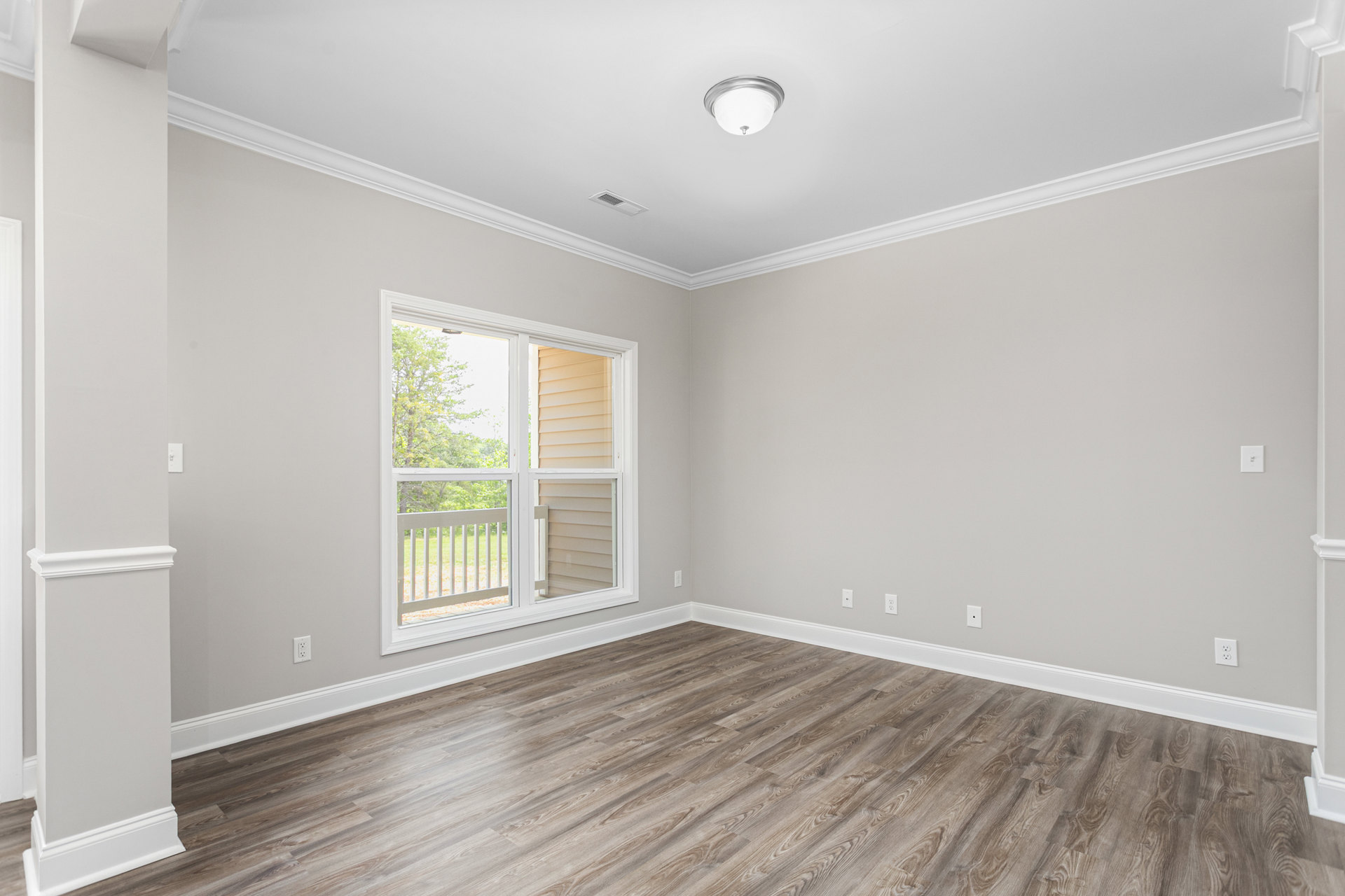 Sunlit room featuring wide-plank wood flooring, white-framed window overlooking balcony with white railing, smooth plaster walls, and decorative ceiling molding.