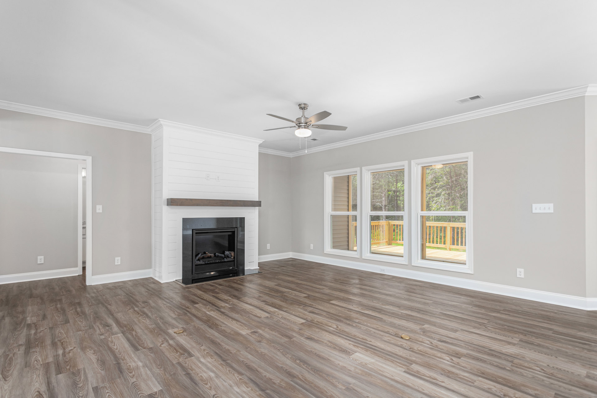 Living room with wood flooring, white walls, built-in fireplace with stacked logs, ceiling fan with light, row of windows overlooking deck and trees.