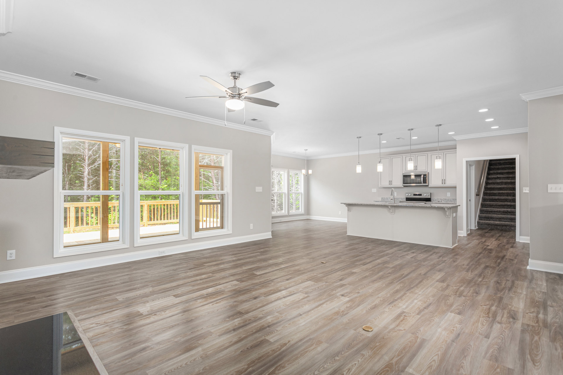 Spacious open floor plan featuring hardwood flooring, ceiling fan with light, white kitchen cabinetry with sink and faucet, built-in microwave, staircase with wood steps, and large