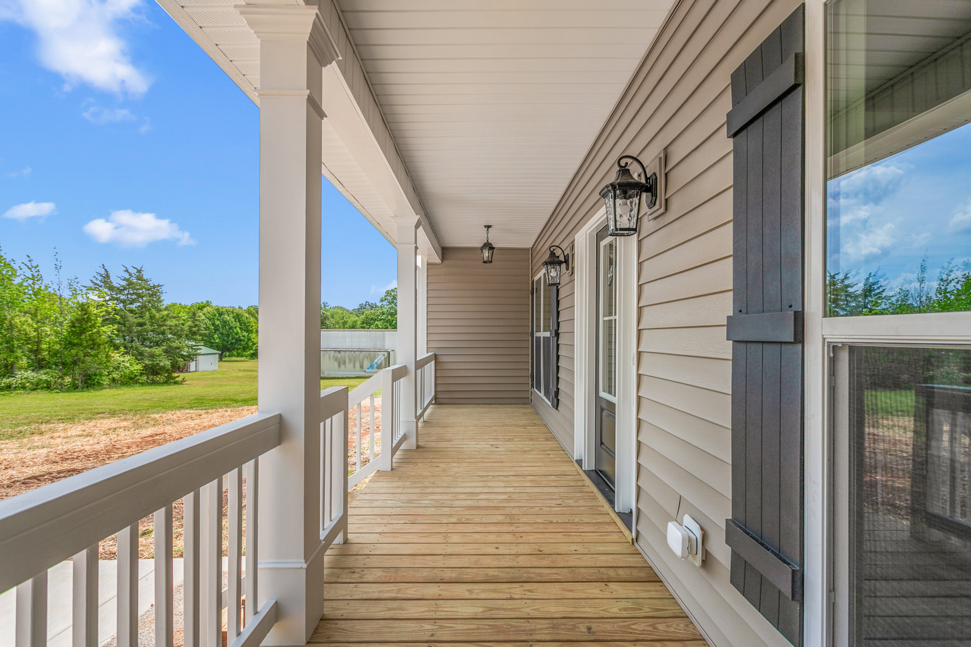 Wood deck porch with white railing, overlooking grass field and trees, light fixture mounted near window reflecting blue sky and clouds