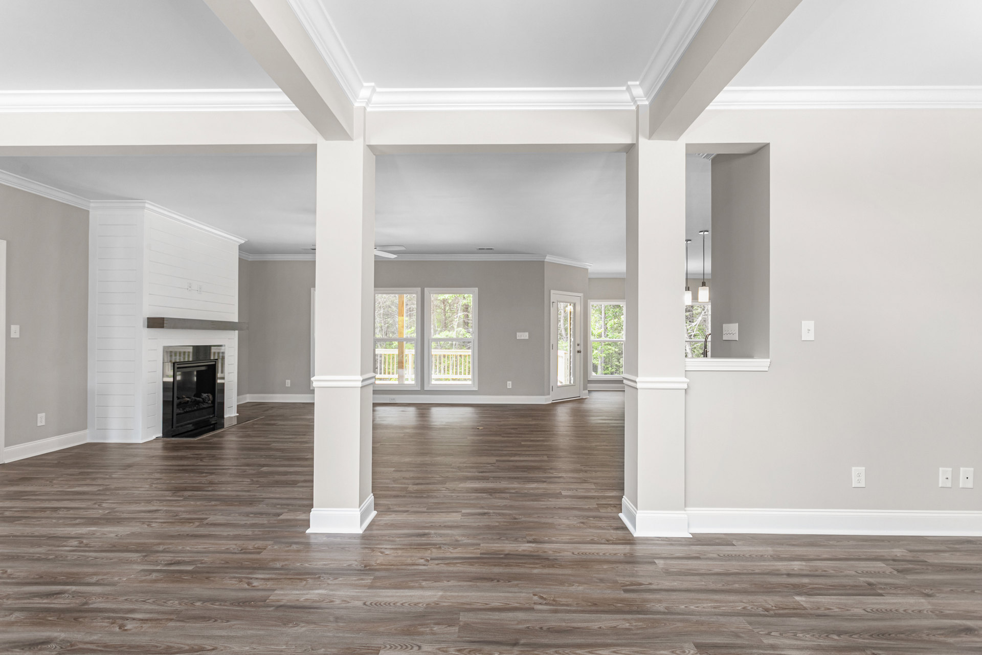 Spacious room with white pillars, wood flooring, black fireplace with glass door, white ceiling with molding, large windows overlooking deck and trees, white walls