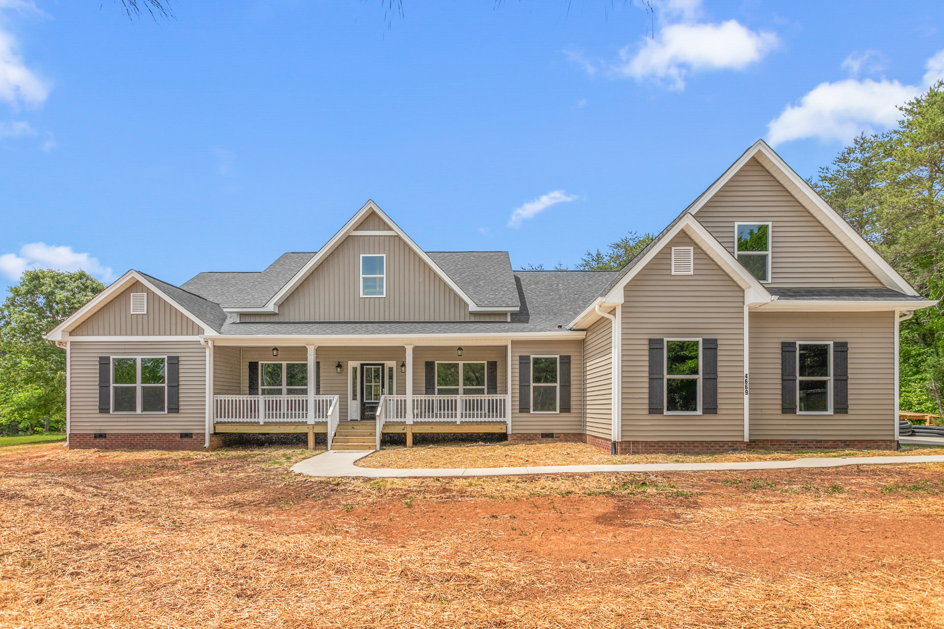 Two-story house with gray roof, white deck railing, multiple windows, dirt and grass driveway, trees and blue sky in the background