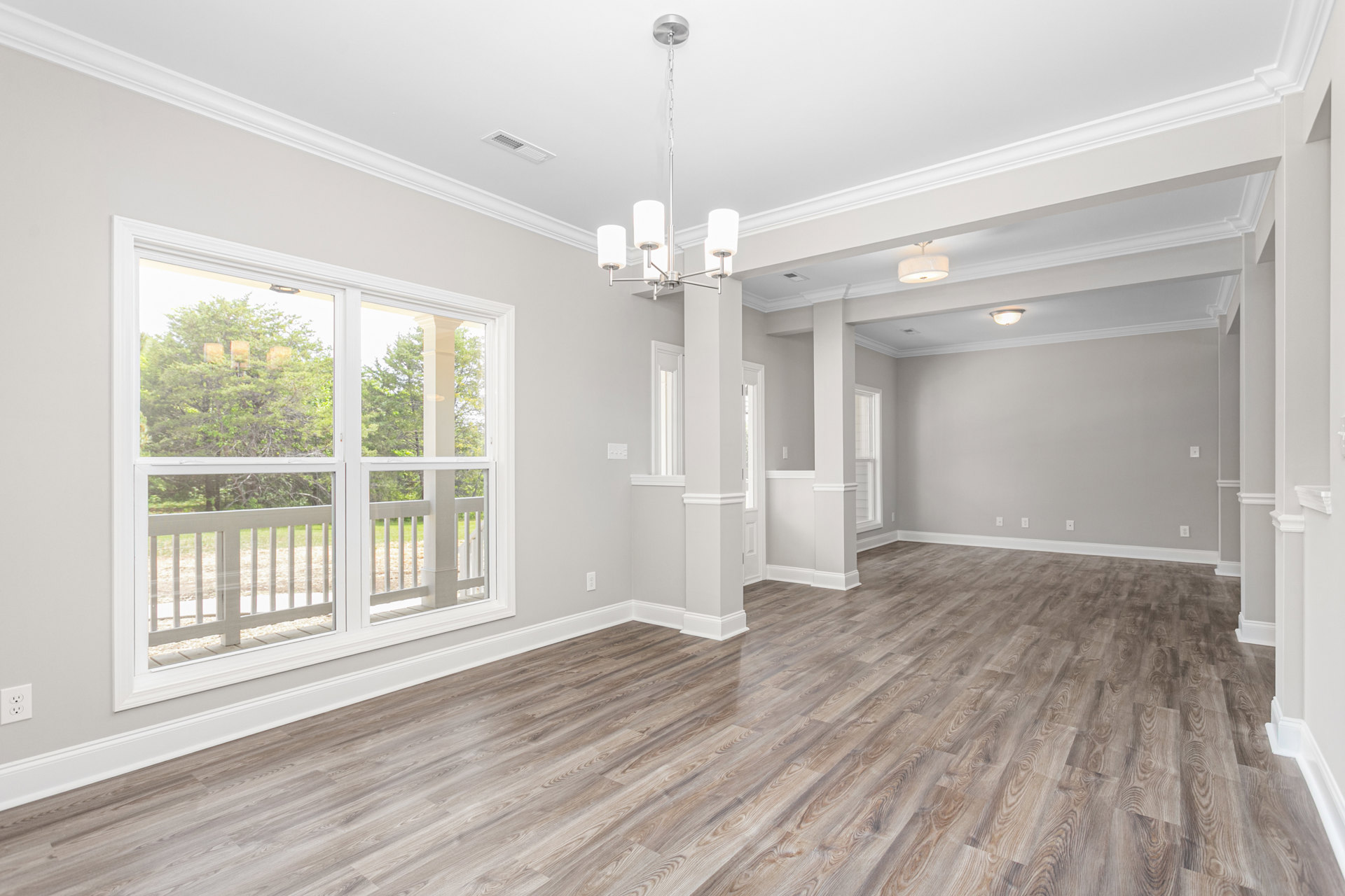 Wood flooring in a spacious room with a decorative chandelier, white window trim, and views of leafy trees outside.