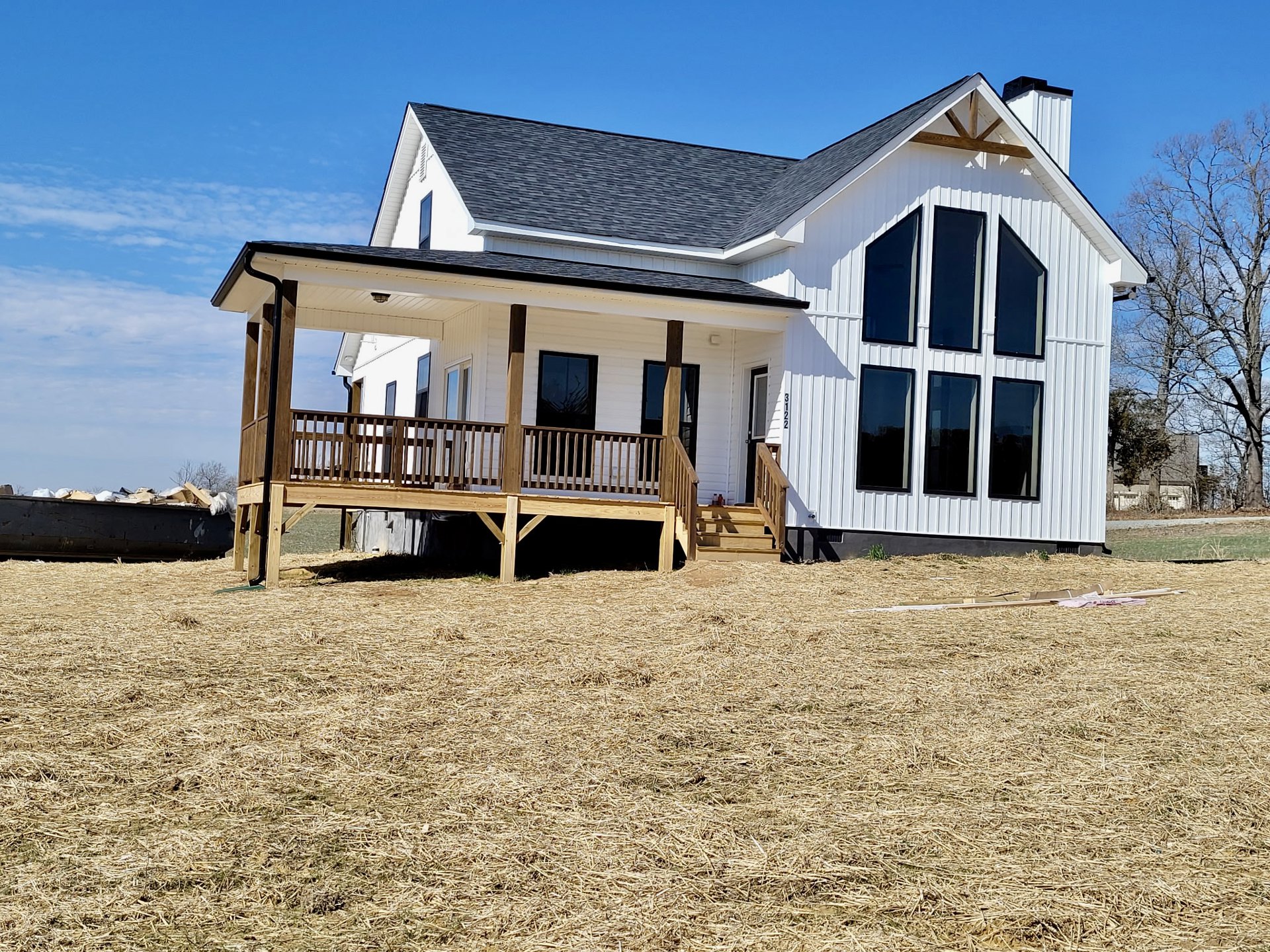 White house with a wooden porch and railing, set on a large grassy hill with a leafless tree nearby, under a partly cloudy sky