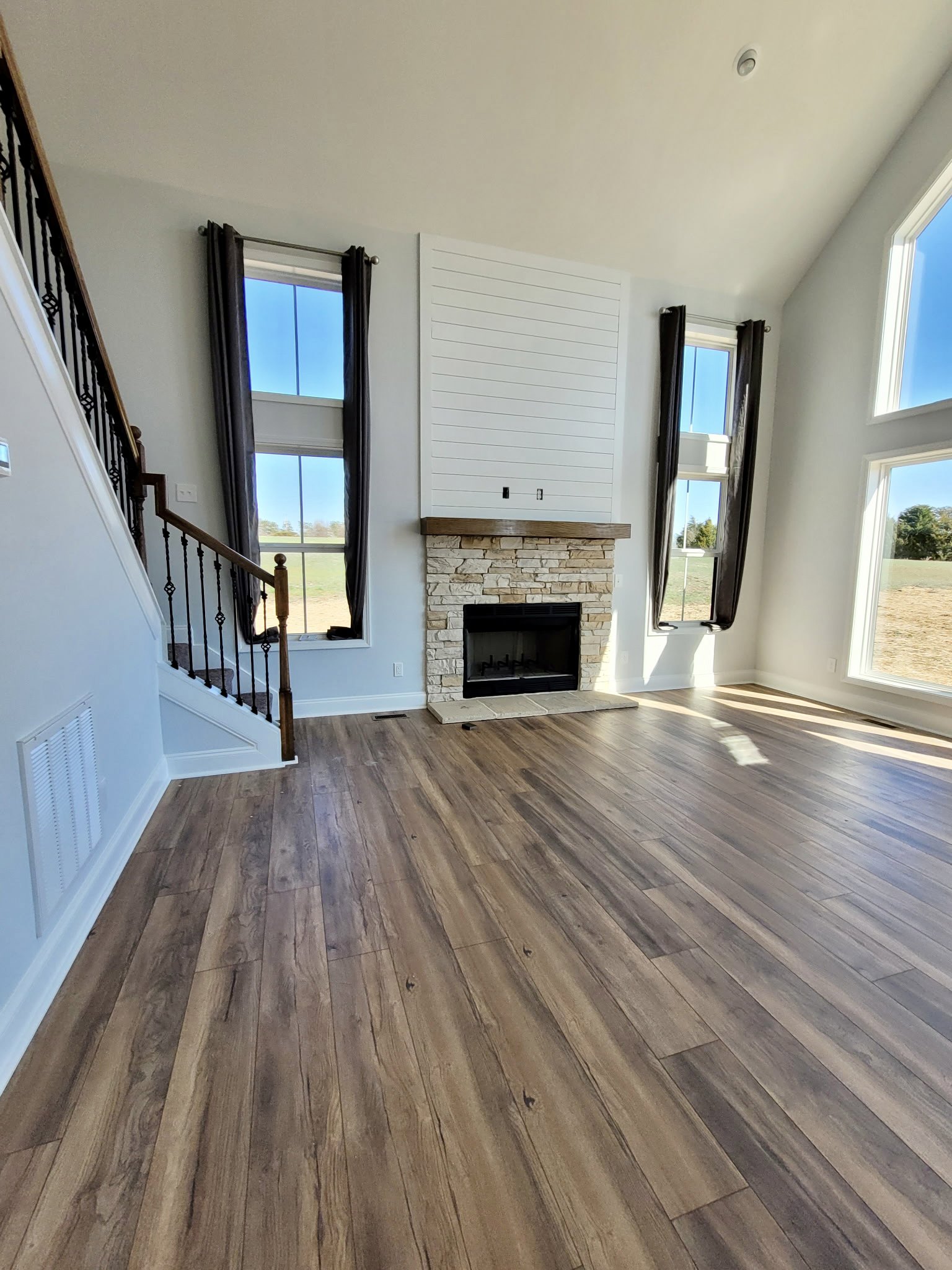 Living room with stone fireplace, wood mantle, hardwood floors, staircase, and large window showing tree and blue sky