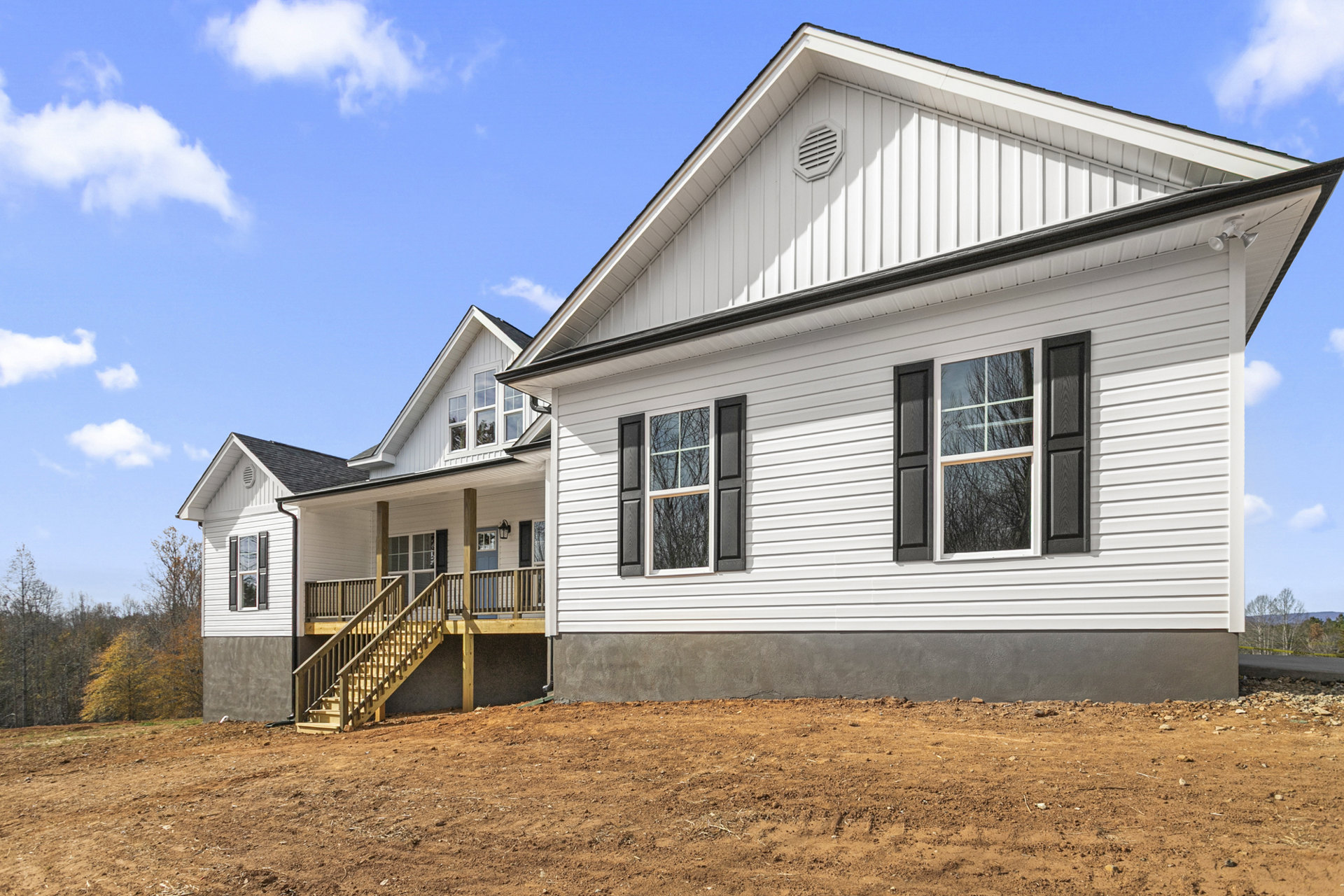 White house with horizontal siding, wooden deck and stairs, large windows reflecting trees, brown dirt field in foreground, white vent on exterior wall, cloudy sky above