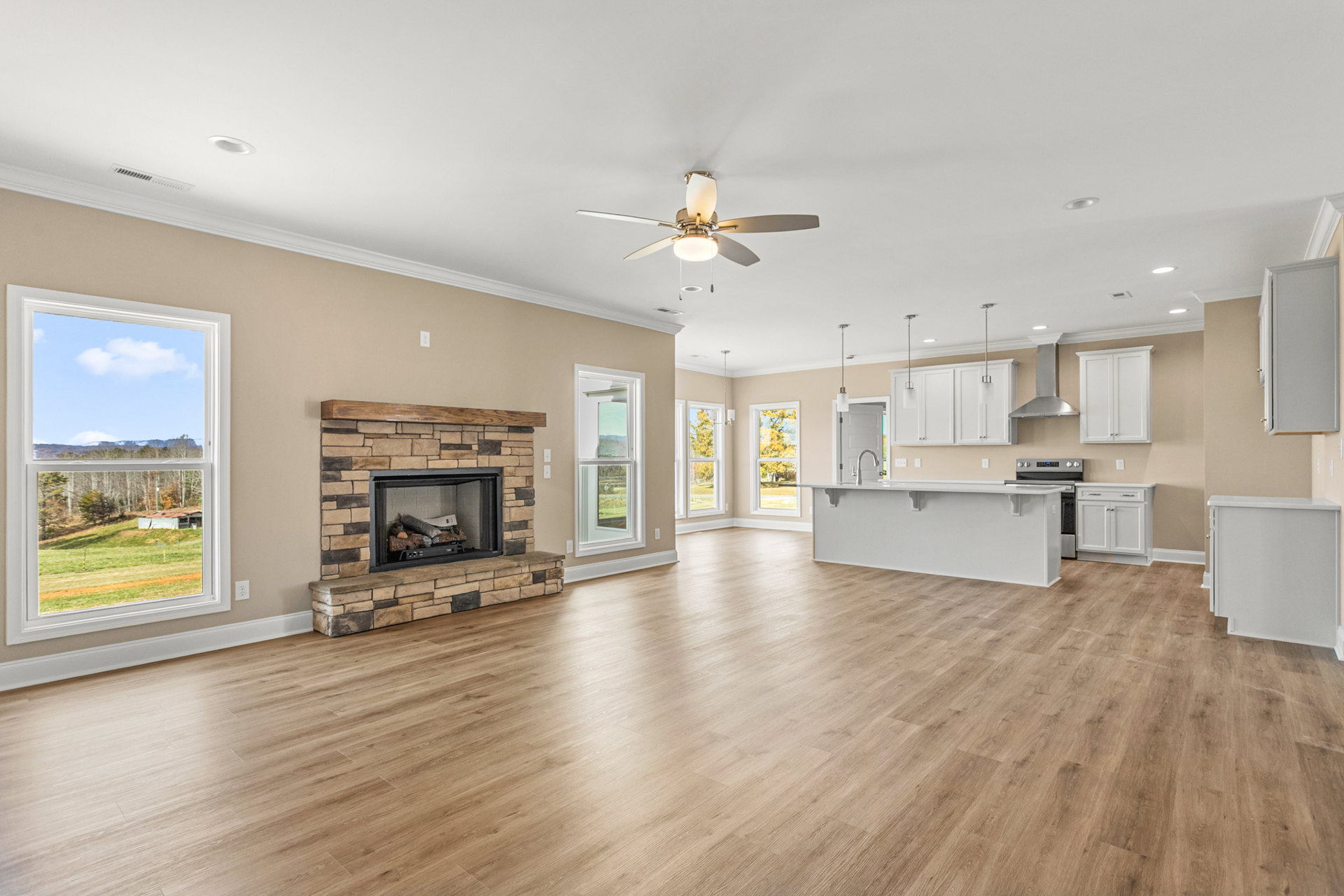 Open concept living room with hardwood floors, stone fireplace filled with wood logs, ceiling fan, and adjacent kitchen; large window offers view of field and trees.