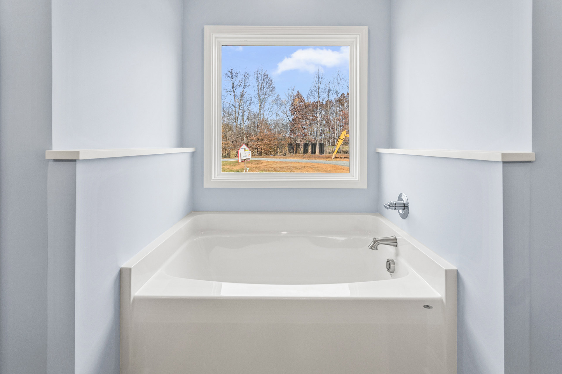 White freestanding bathtub beneath a large window with blue accent wall, chrome faucet, playground and yellow construction equipment visible outside