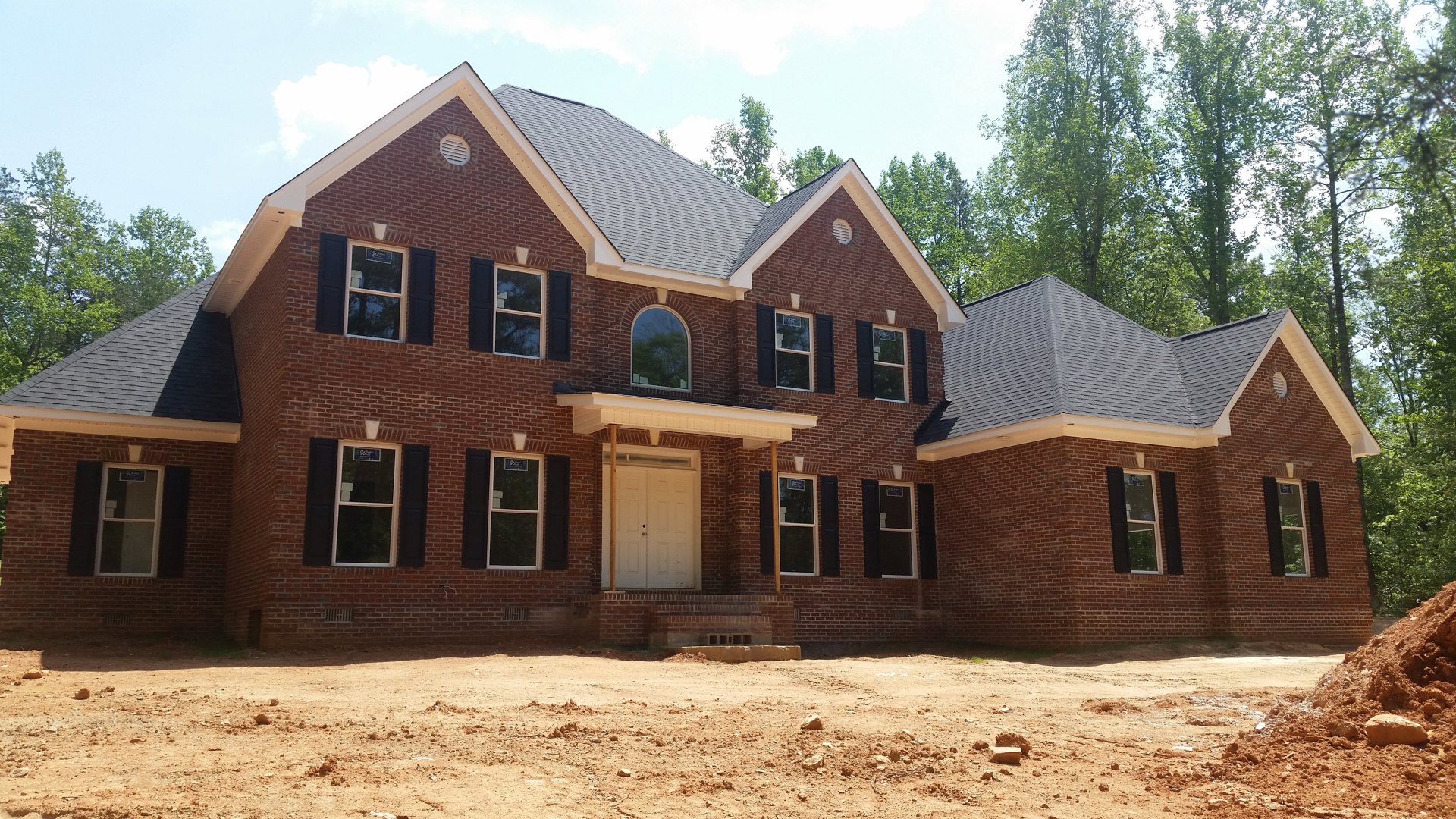 Red brick house with white double doors and white-framed windows, dirt yard in front, tree visible through window, clear sky overhead