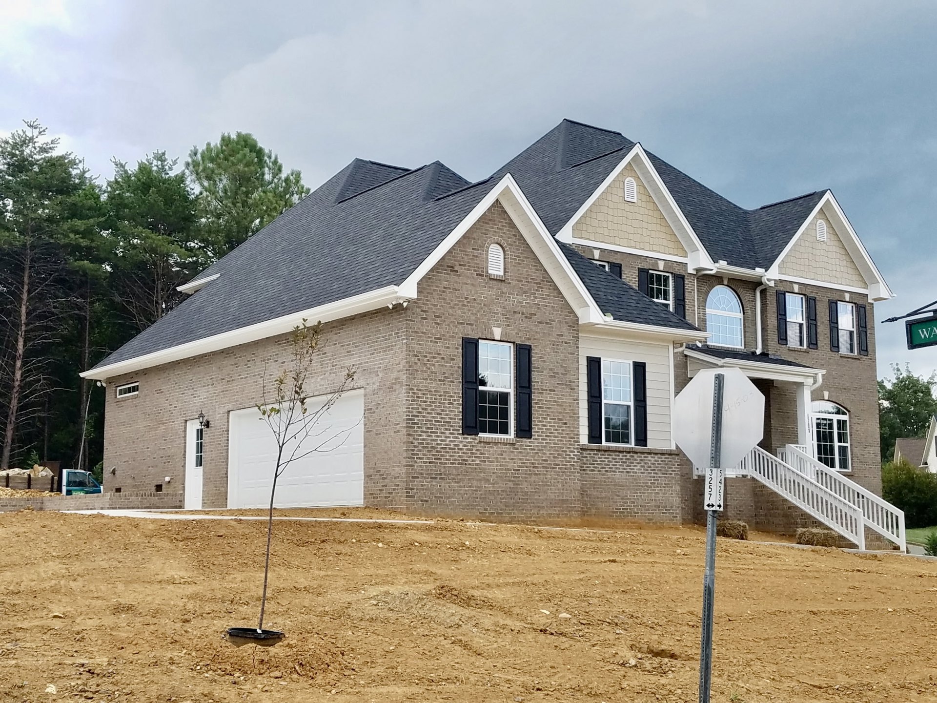 Brick house with white-framed windows, large paved driveway, white staircase railing, tree near garage door, stop sign on pole, partly cloudy sky