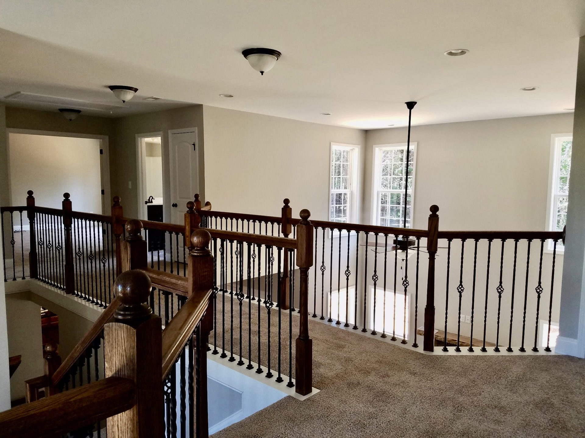Brown carpeted staircase with wooden railing, white baluster, and white-framed window overlooking trees; ceiling-mounted light fixture with white shade.