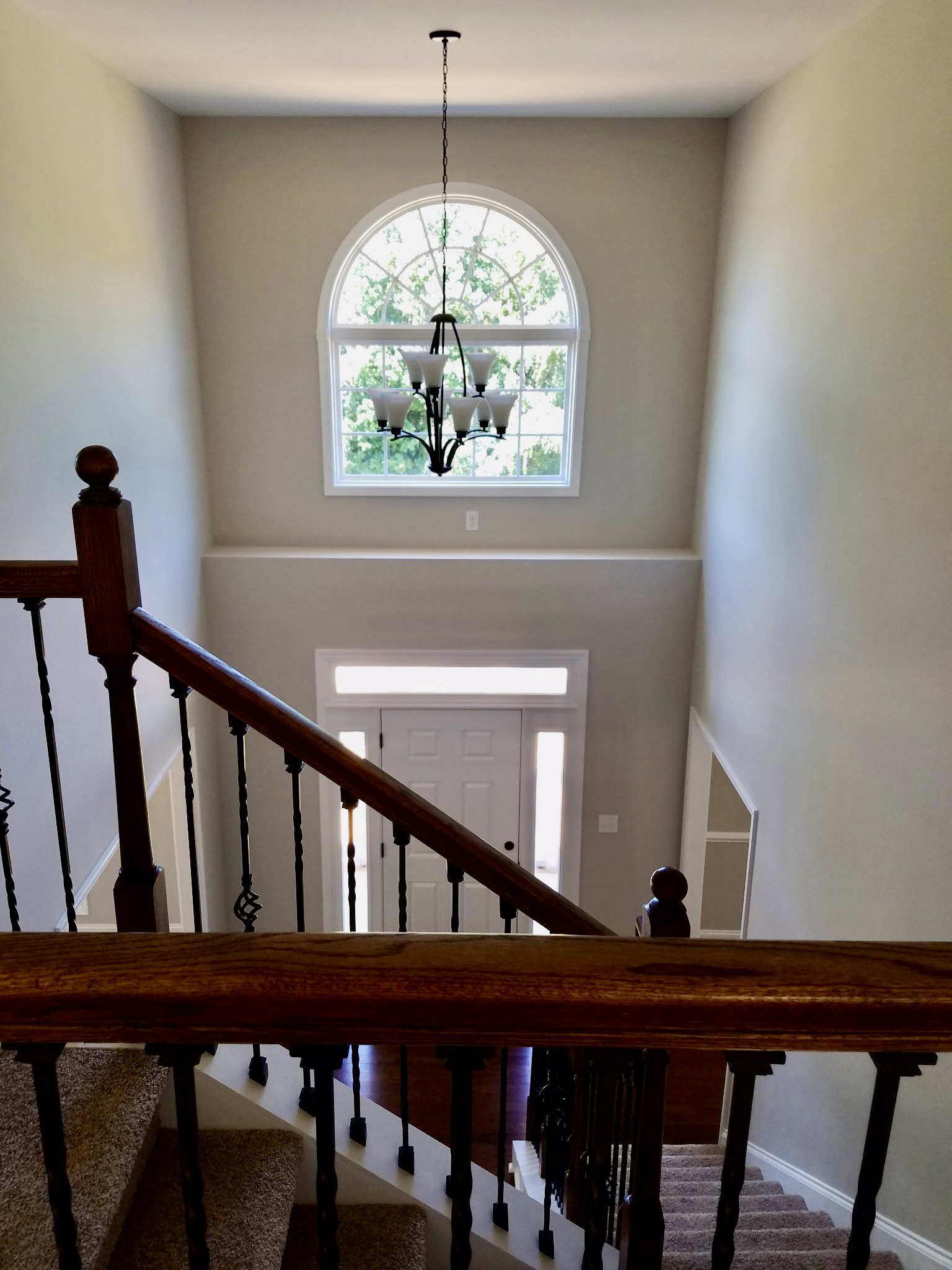 Wooden staircase with white balusters, modern chandelier hanging from ceiling, large window providing natural light, white door adjacent to stairway.