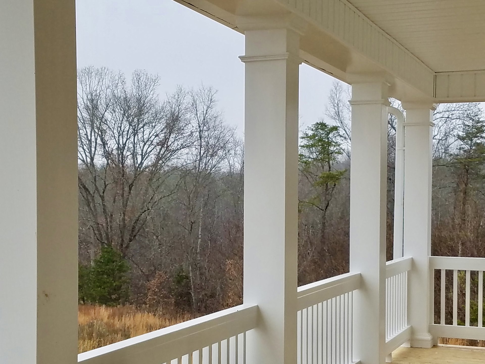 Front porch with white pillars, white railing, concrete floor, and green-leaved trees outside