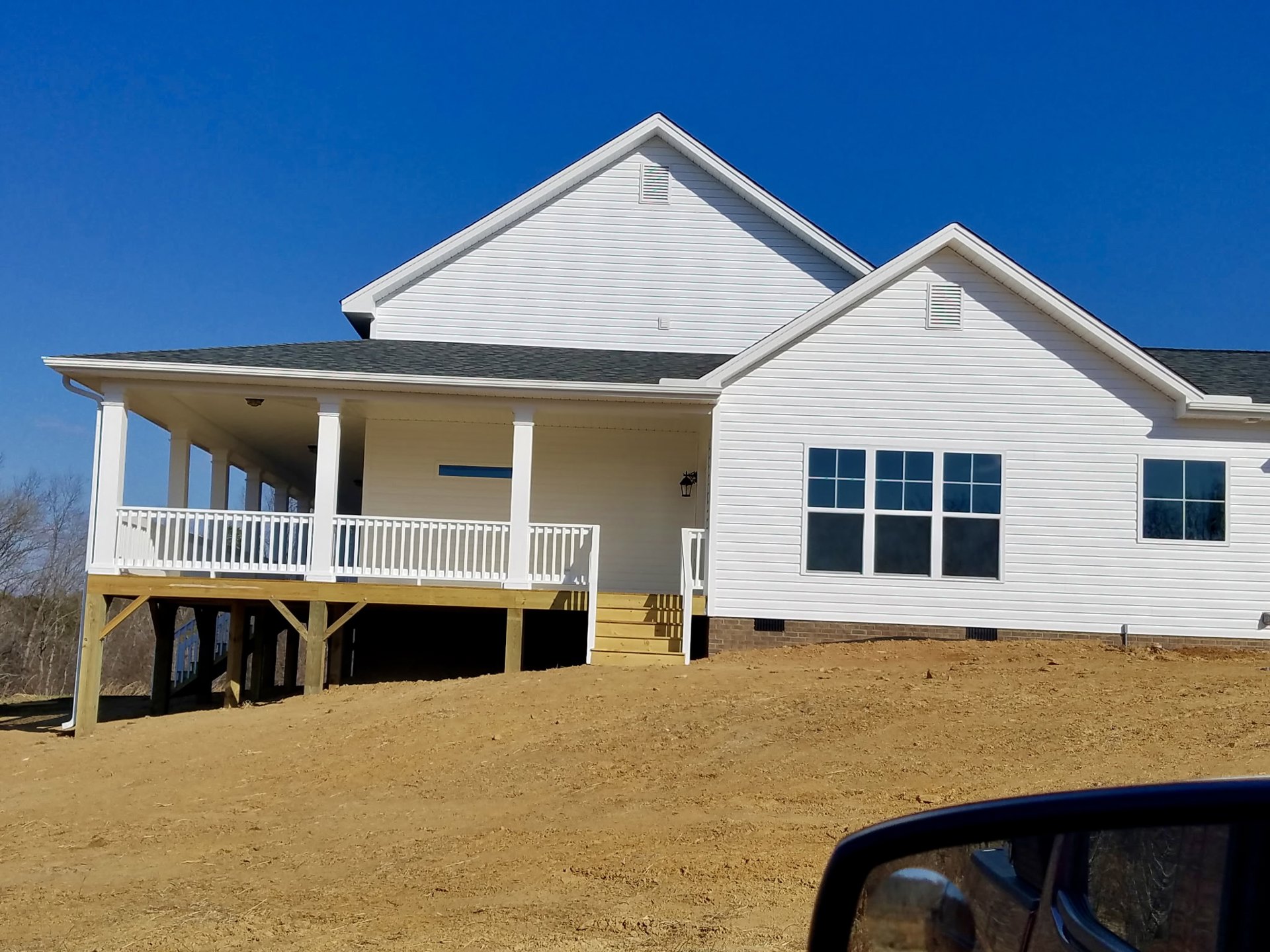 White house with covered porch and wooden deck, white railings, large windows, Southfork Ranch visible in the background under blue sky.
