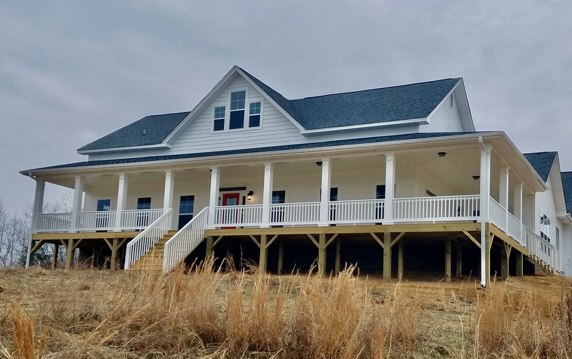 White siding house with blue roof, red front door, covered porch with white railings, tall grass in front, multiple windows, tree branches dusted with snow.