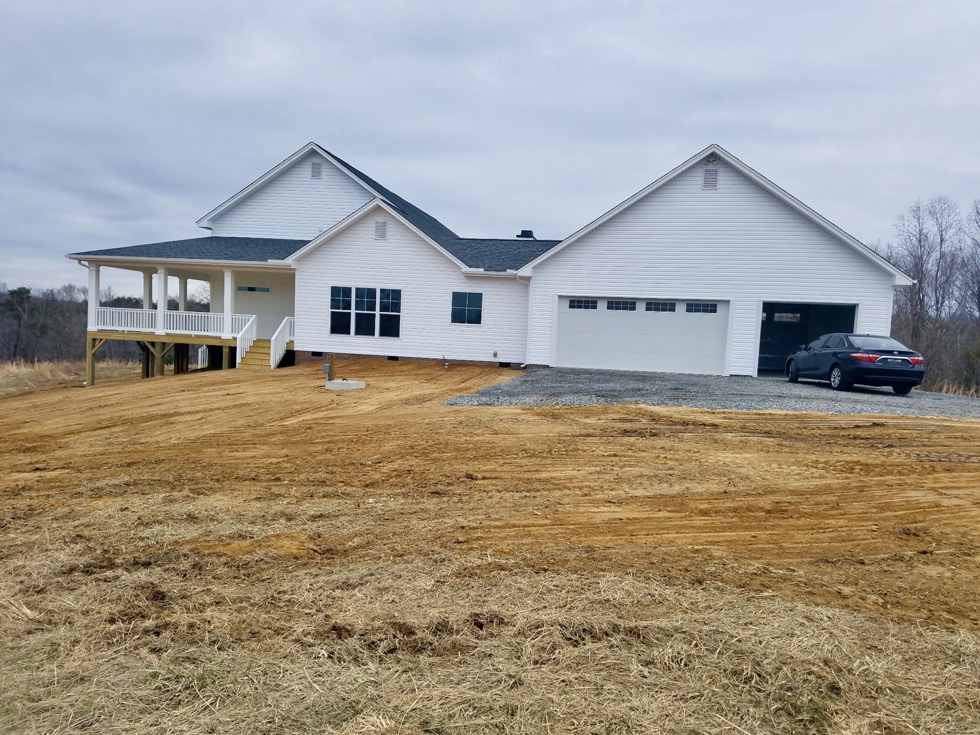 Two-story house with white siding, attached garage, black car parked on concrete driveway, wooden porch with white railing, four-pane window, grassy yard, trees and cloudy sky in