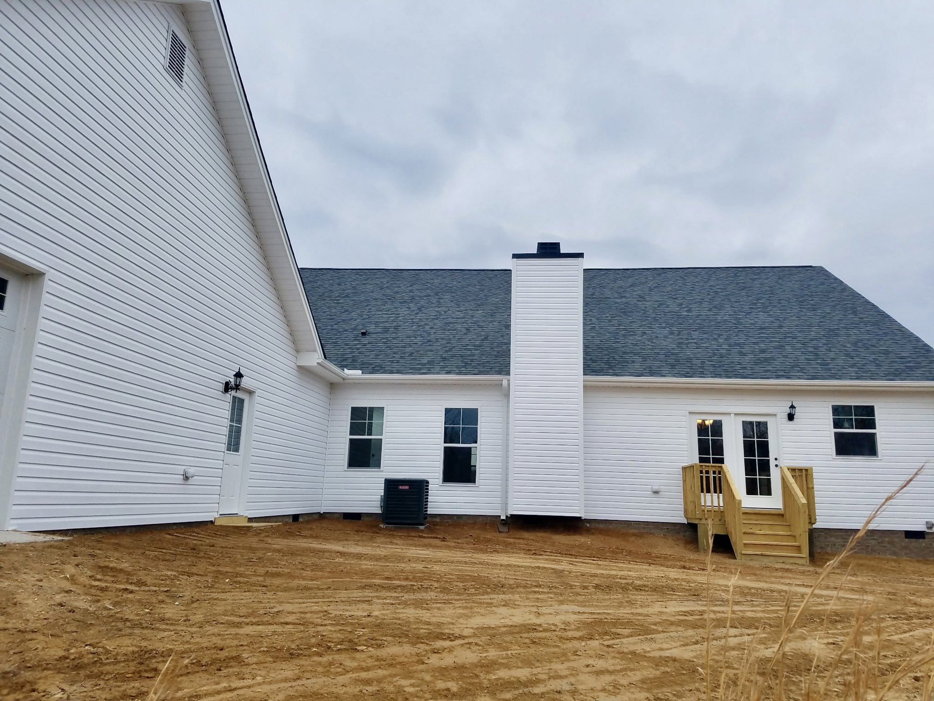 Modern house with white vertical siding, black-framed window, wooden stairs leading to entry door, chimney on roof, dirt yard in foreground, black square object with red label near