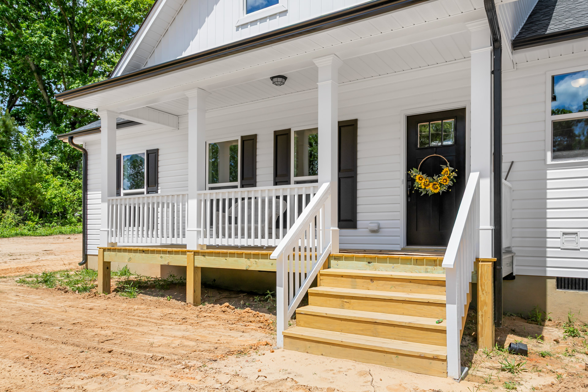 Two-story home with white siding, wooden porch stairs, white railing, sunflower wreath on front door, sandy ground, and leafy trees in background