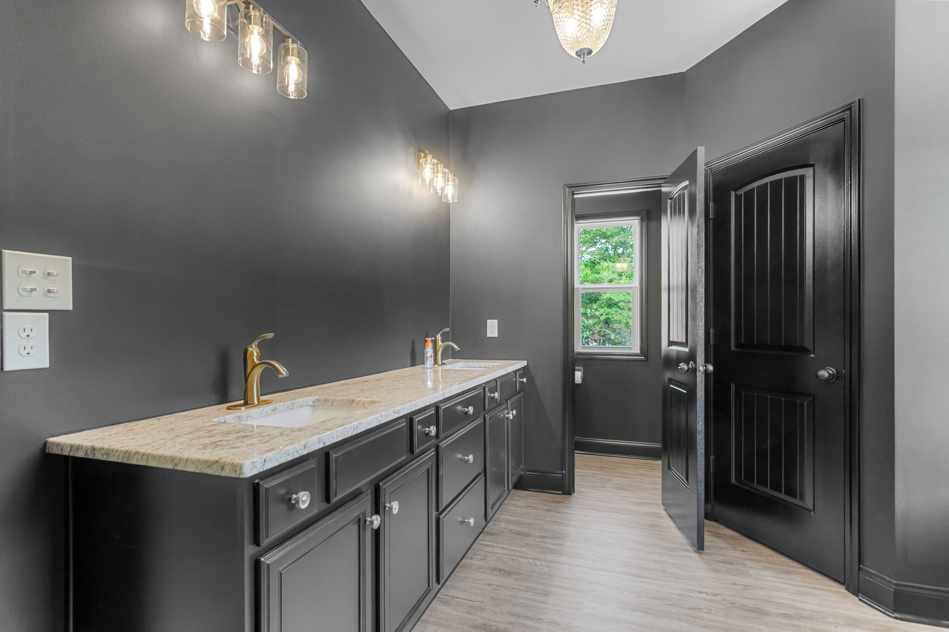 Bathroom with double white sinks on a stone countertop, dark cabinetry, chrome faucets, crystal chandelier, black door with silver knobs, window overlooking trees, light fixture