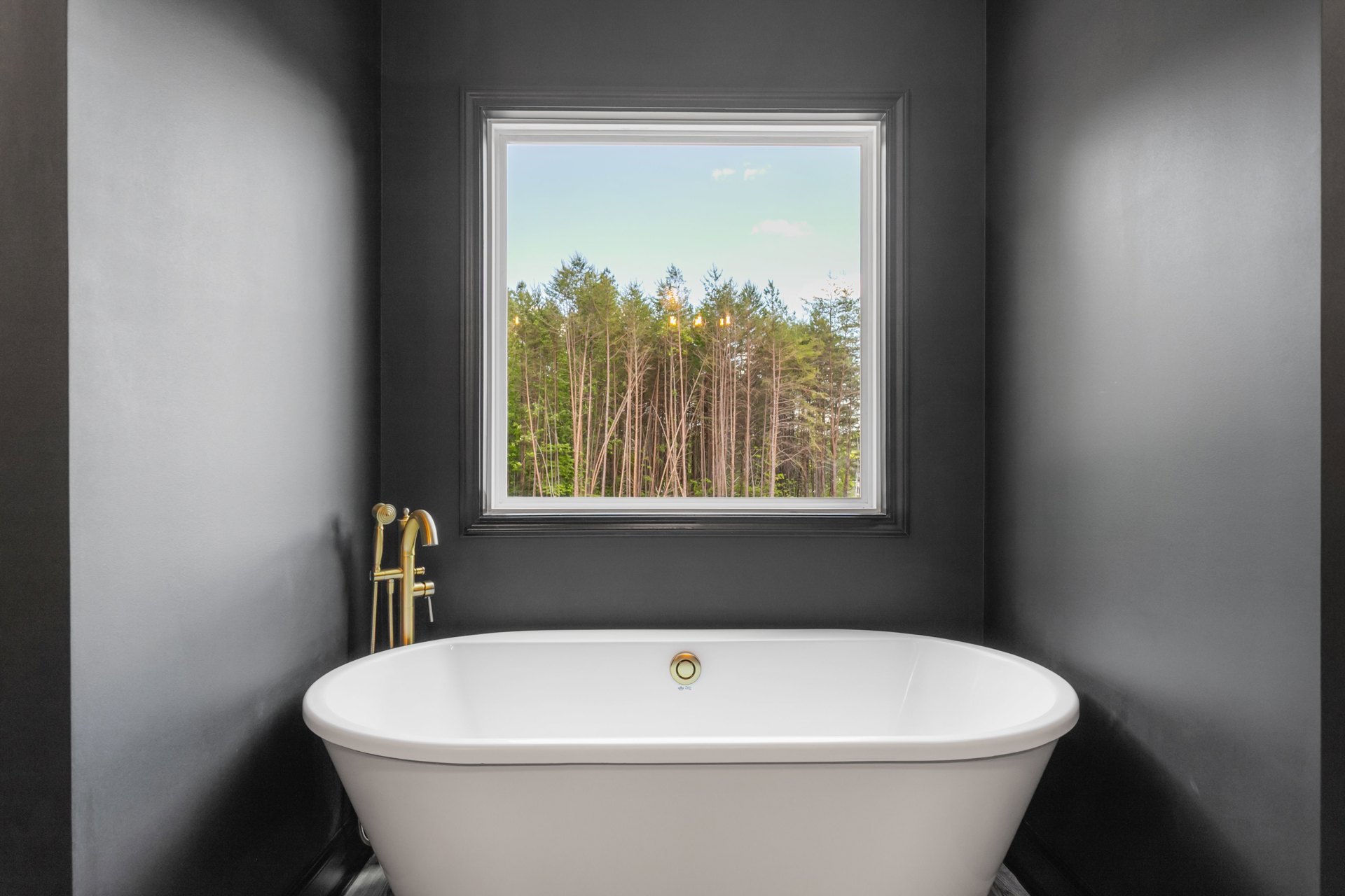 Freestanding white bathtub beneath large window with tree views, gold faucet and handheld shower, light gray walls, soft natural light, subtle shadow on wall