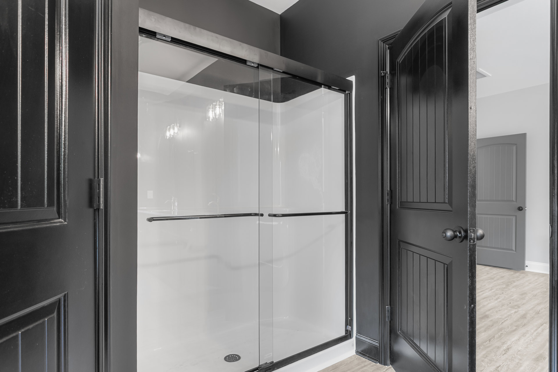 Glass shower enclosure with black metal handles, white tile walls, and light wood flooring