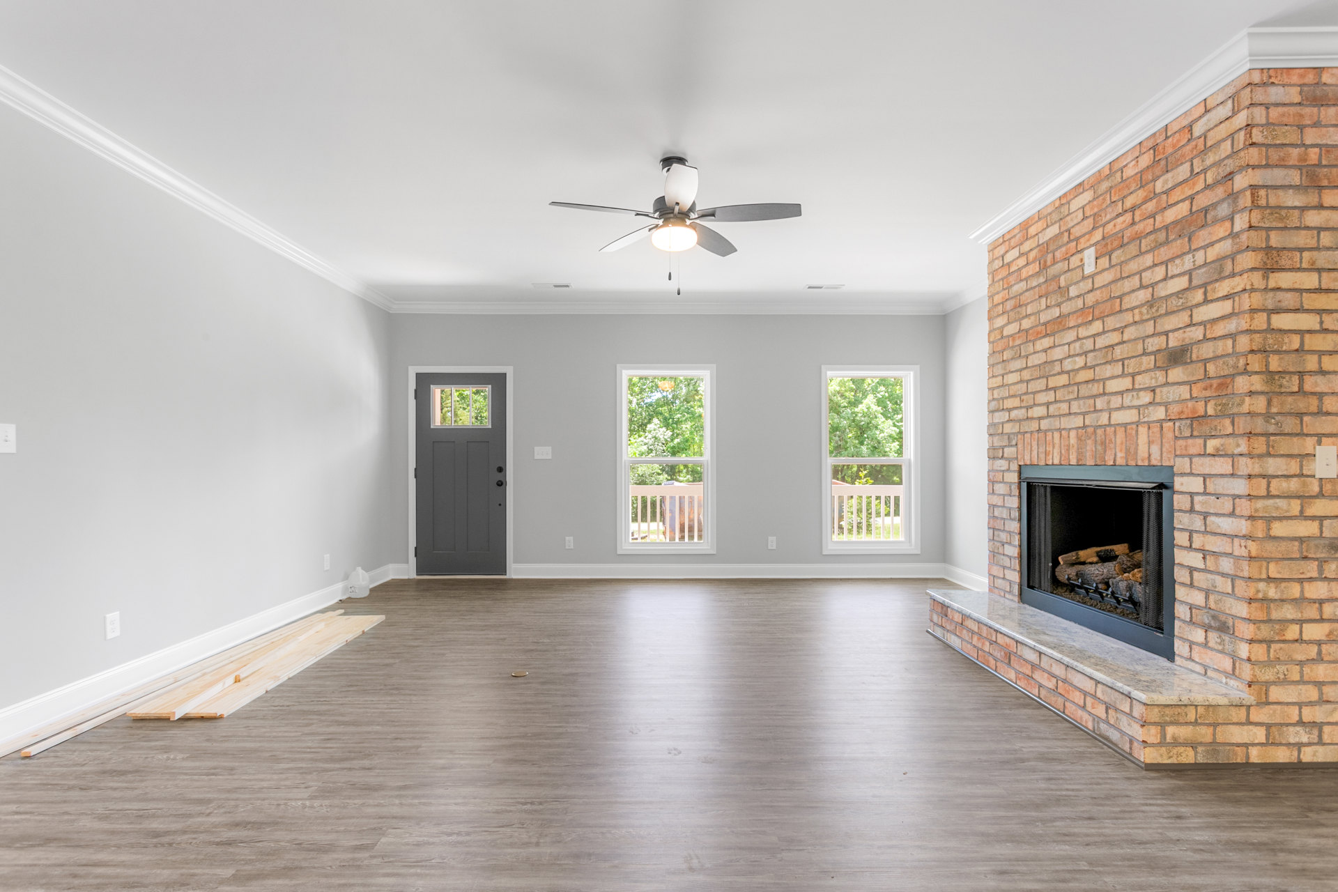 Living room featuring a brick fireplace with stacked wood, ceiling fan with light fixture, large windows showing trees outside, and a grey door with glass panel.