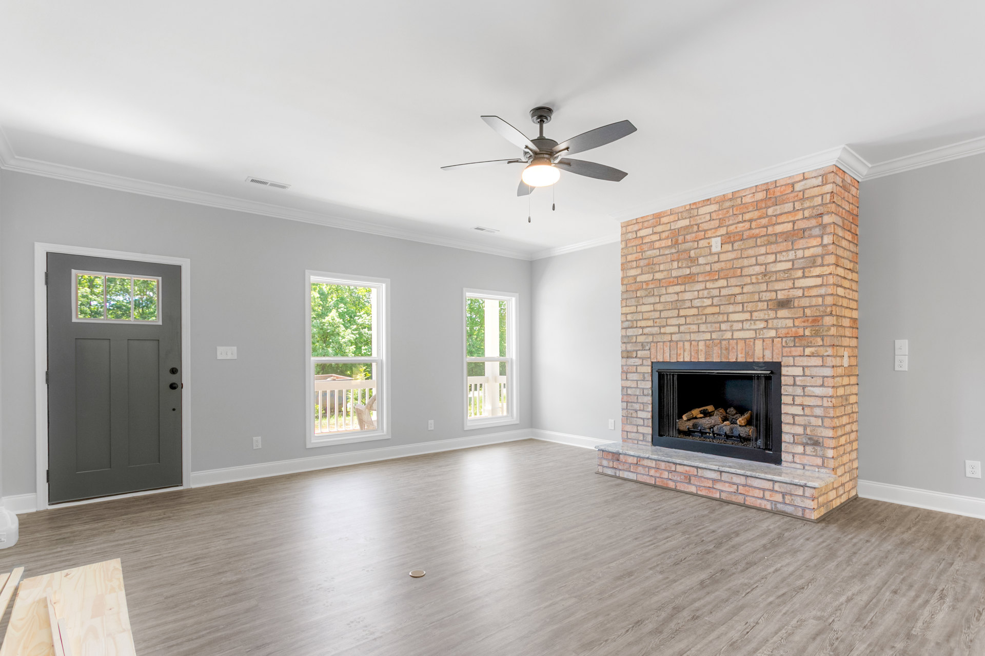 Living room featuring a brick fireplace with stacked logs, wooden flooring, ceiling fan with light fixture, grey door with window, and large window overlooking trees.