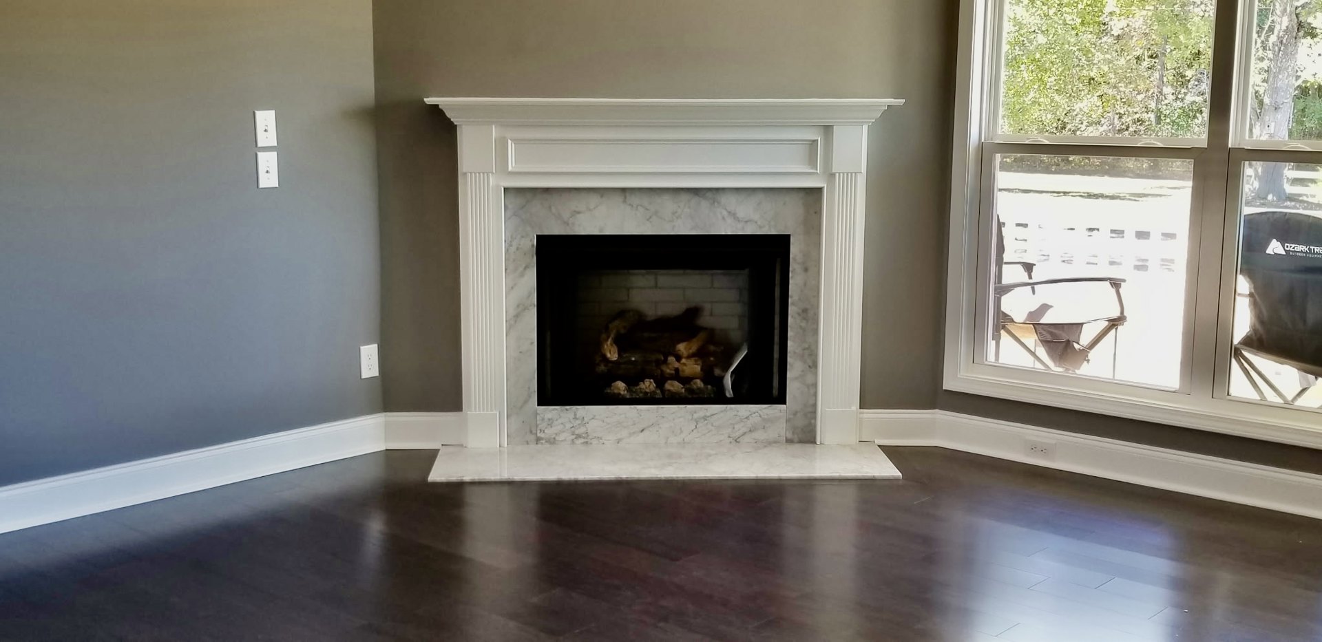 White-framed fireplace with stacked firewood, dark wood flooring with white trim, large window showing outdoor chair, white wall outlet and key on white wall