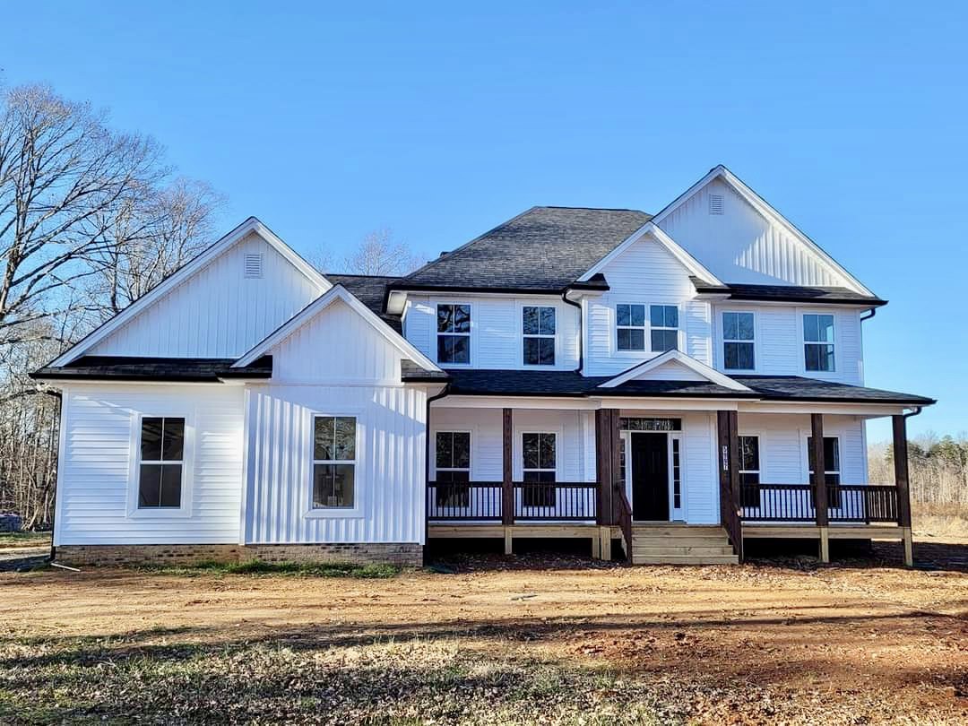 White two-story house with black door and white trim, covered porch, white-framed windows, brick wall, dirt path bordered by grass, and gray shingle roof under a partly cloudy sky.