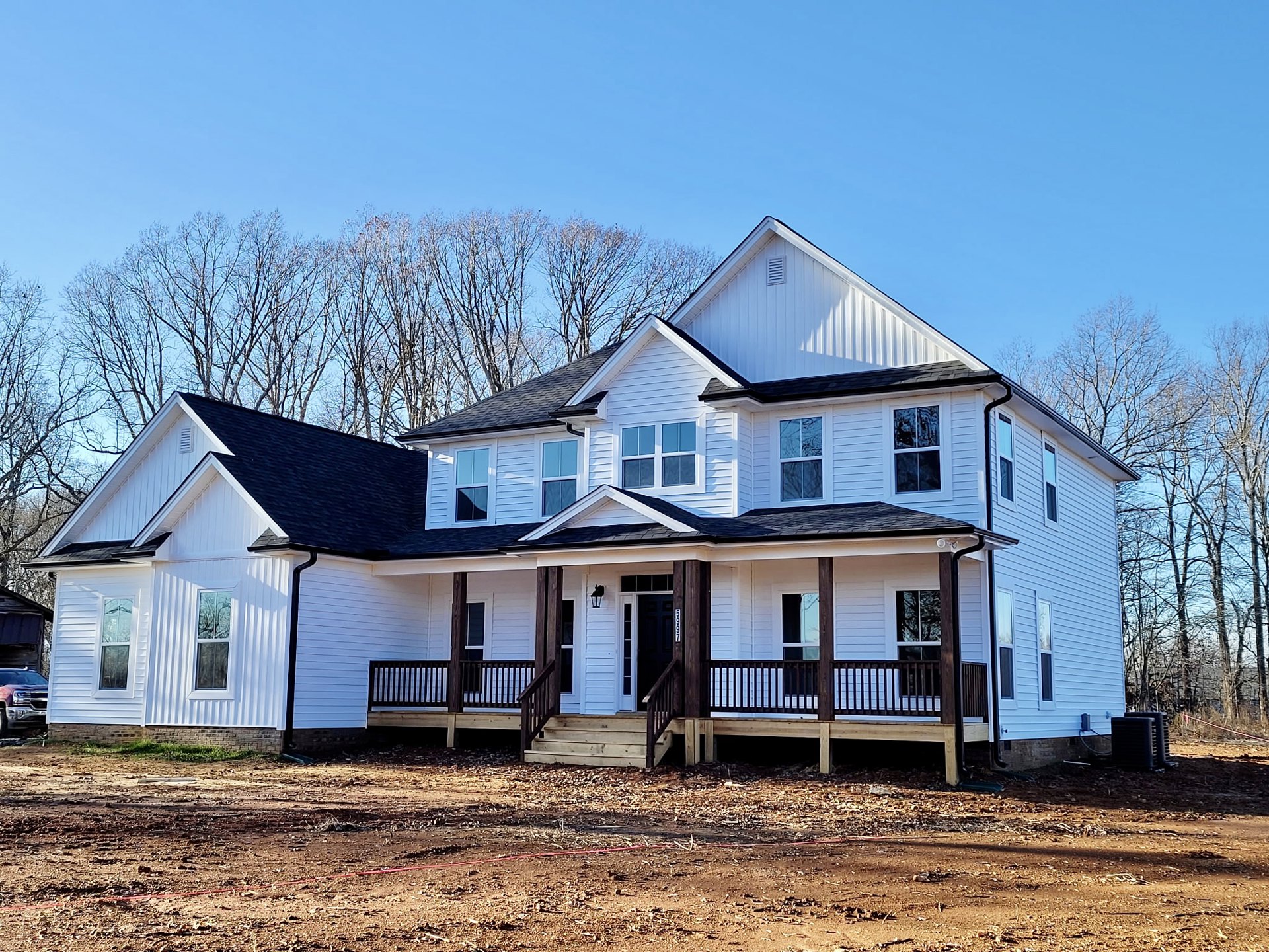 White two-story house with black gabled roof, covered front porch, wooden stairs, large windows reflecting nearby trees, and a parked car in the driveway