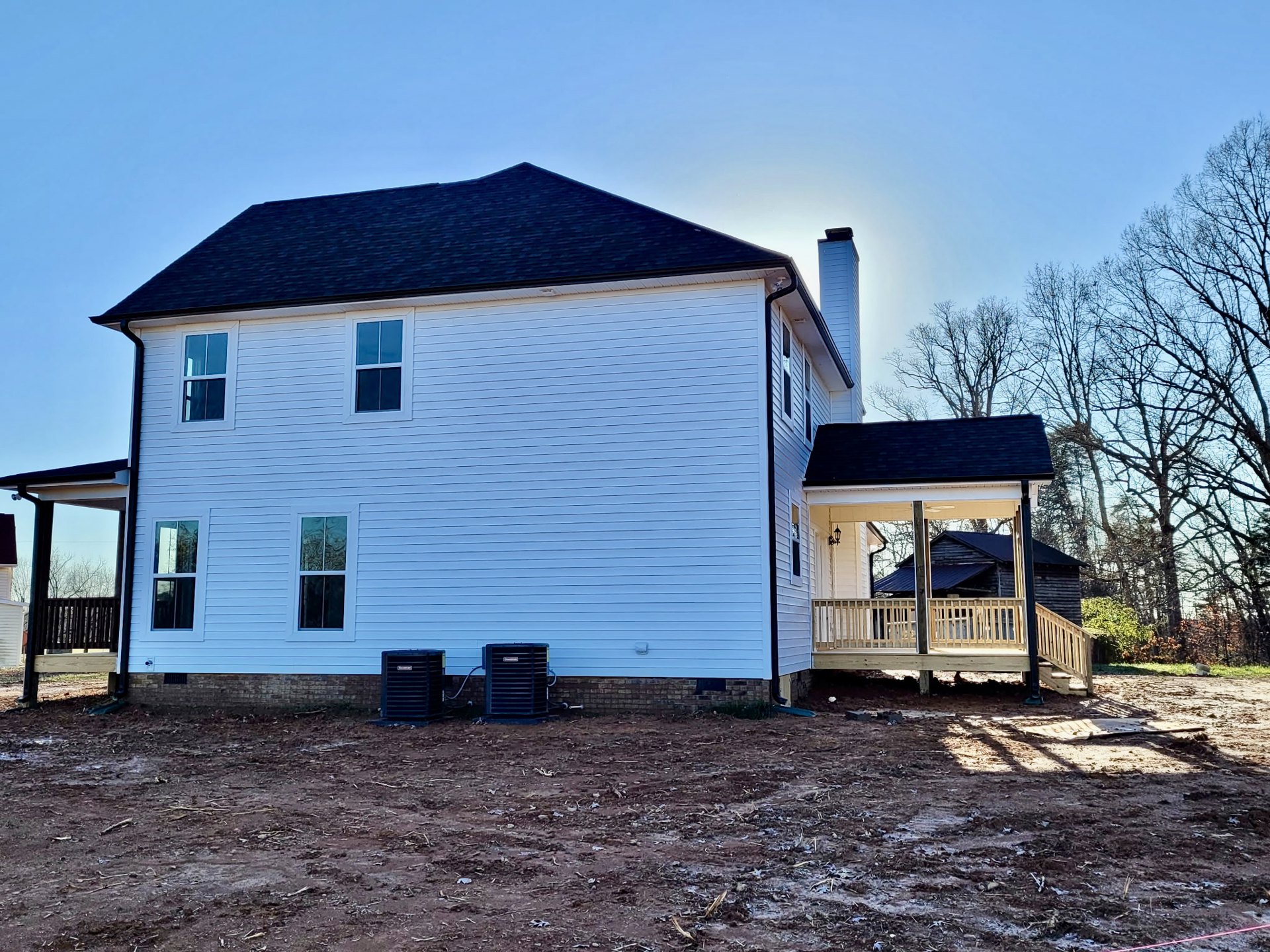 White house with black roof, front porch with white railings, elevated wooden deck, multiple white-framed windows, dirt yard with black metal vent object, trees in background.